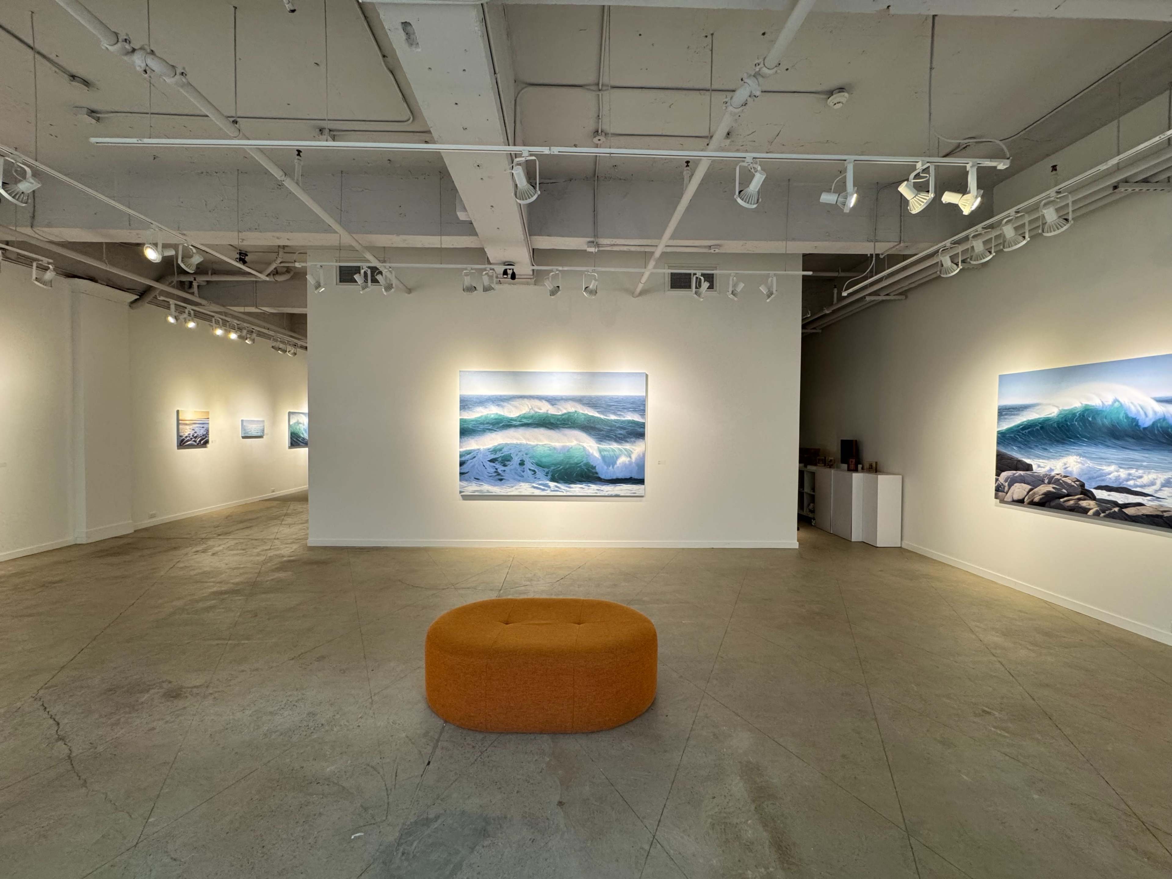 The image shows a modern art gallery featuring several large wave paintings on white walls and a circular orange ottoman in the center of a concrete floor.