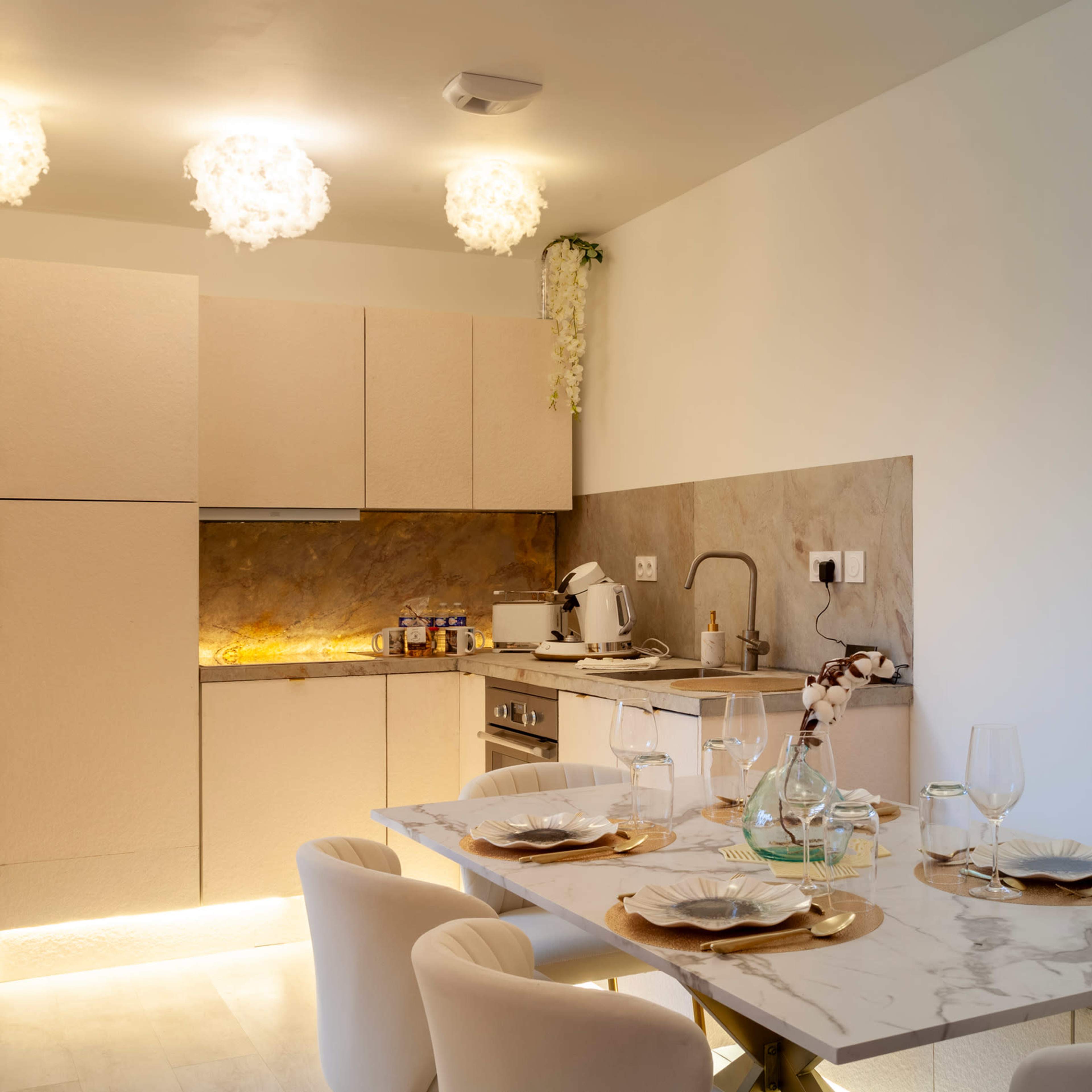 A modern kitchen features beige cabinetry, a marble countertop, a dining table set with glassware and plates, and soft lighting from decorative fixtures.