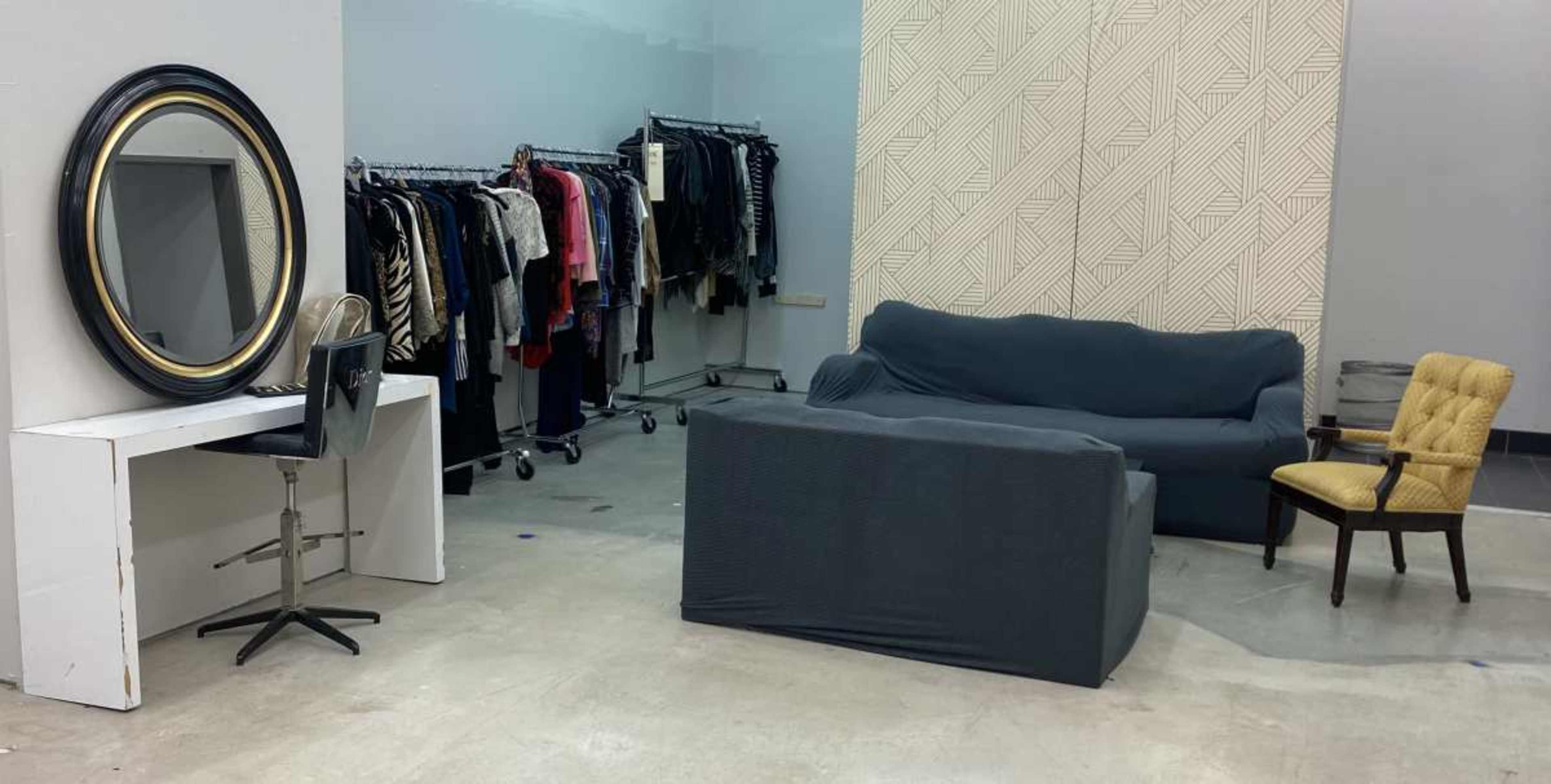 The image shows a room with clothing racks on one side, a mirror and desk on another, a gray sofa, and a yellow chair.