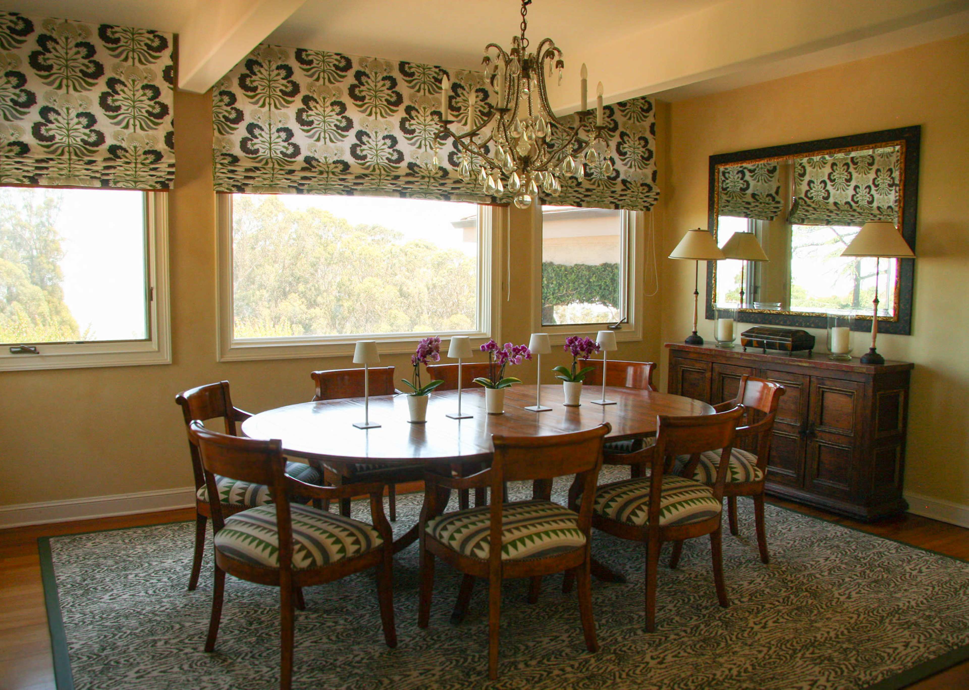 A circular wooden dining table surrounded by eight chairs is set in a well-lit room with large windows, a chandelier, and a sideboard.