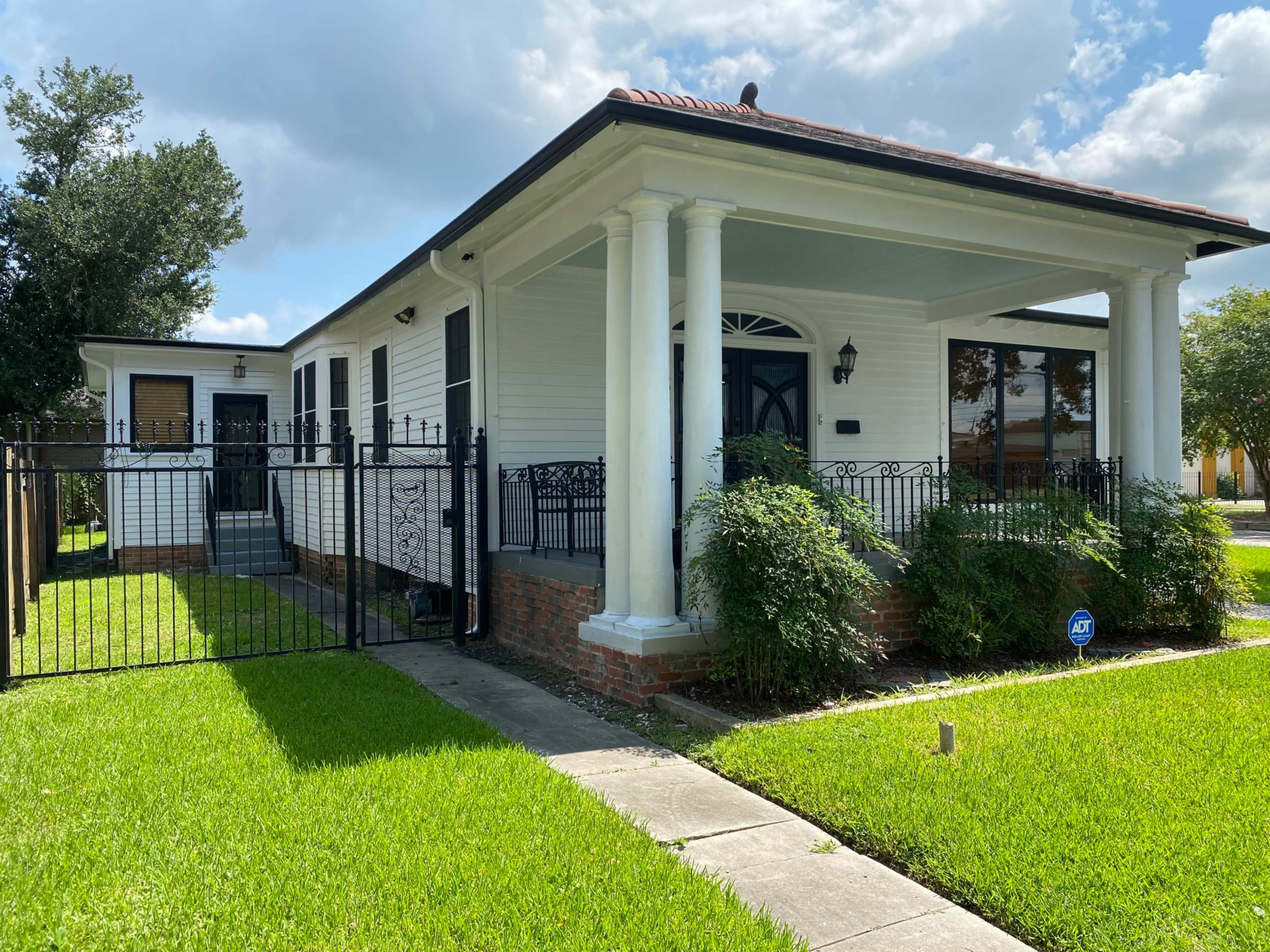 The image shows a white, single-story house with a red-tiled roof, surrounded by a black wrought iron fence and a well-manicured lawn.