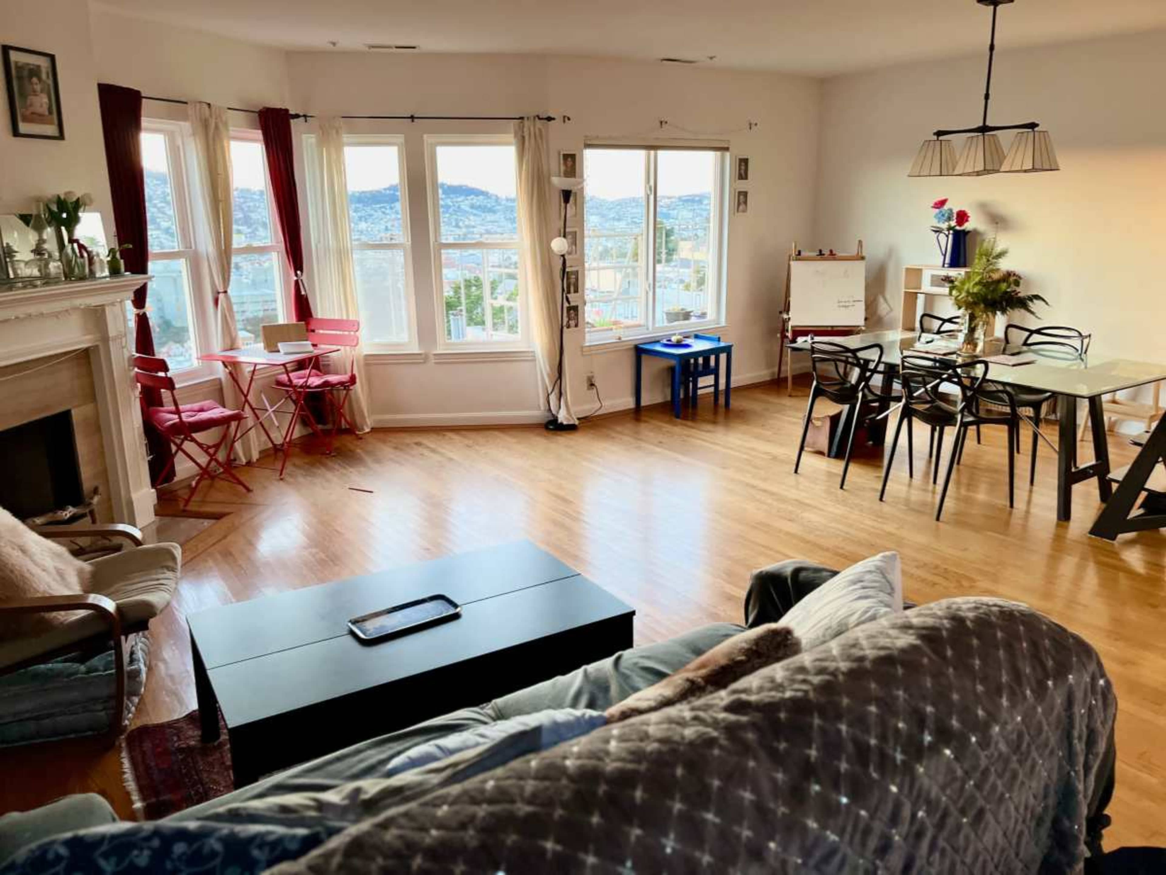 The scene shows a bright living room with wooden flooring, large windows, and a mix of modern and cozy furniture, including a sofa, dining table, and chairs.