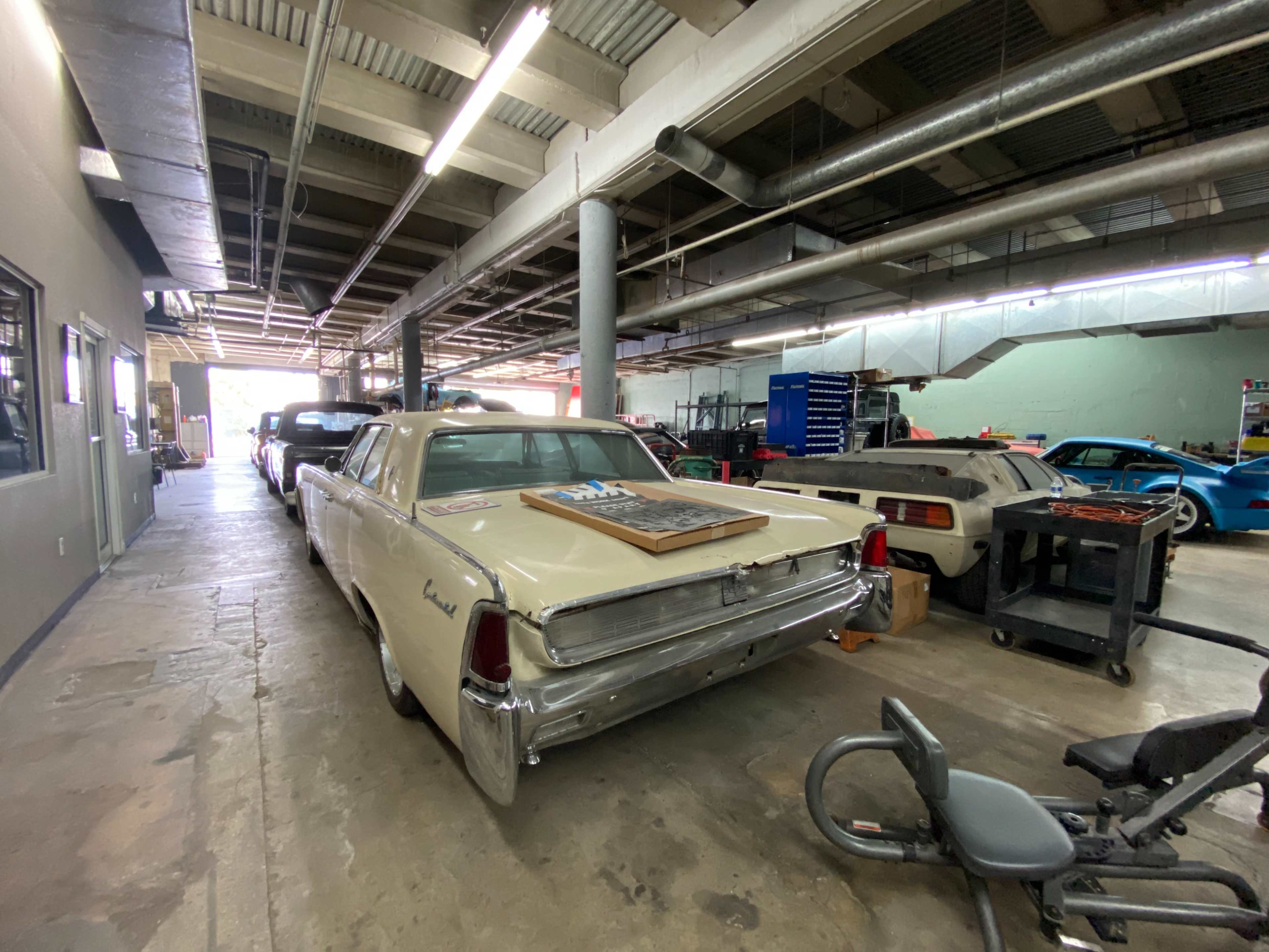 The image shows a garage filled with various vehicles, including a classic beige car in the foreground and several other cars and equipment in the background.