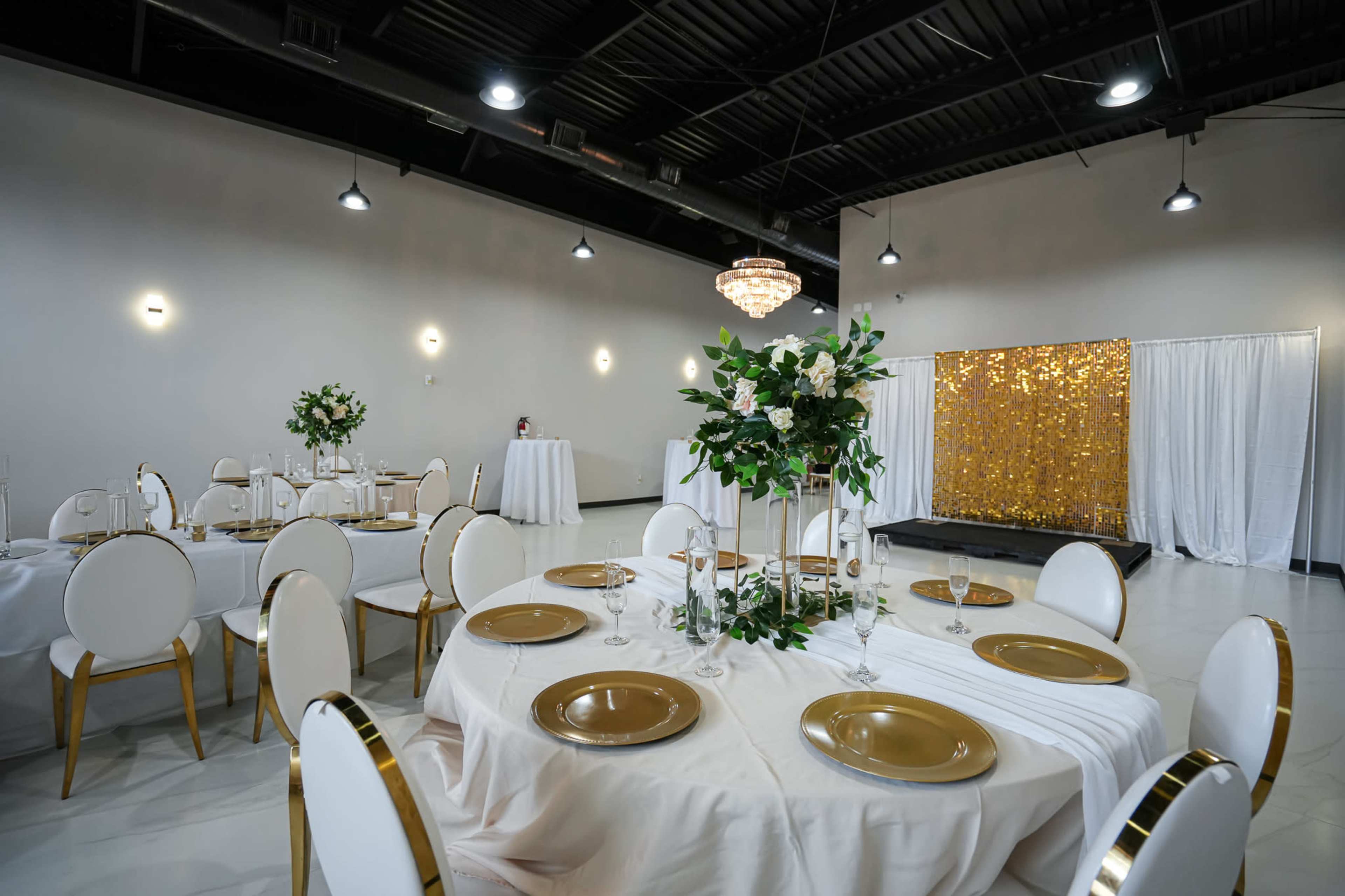 A dining area is set up with round tables adorned in white linens and gold accents, featuring floral centerpieces, with a backdrop of a glittering gold curtain.