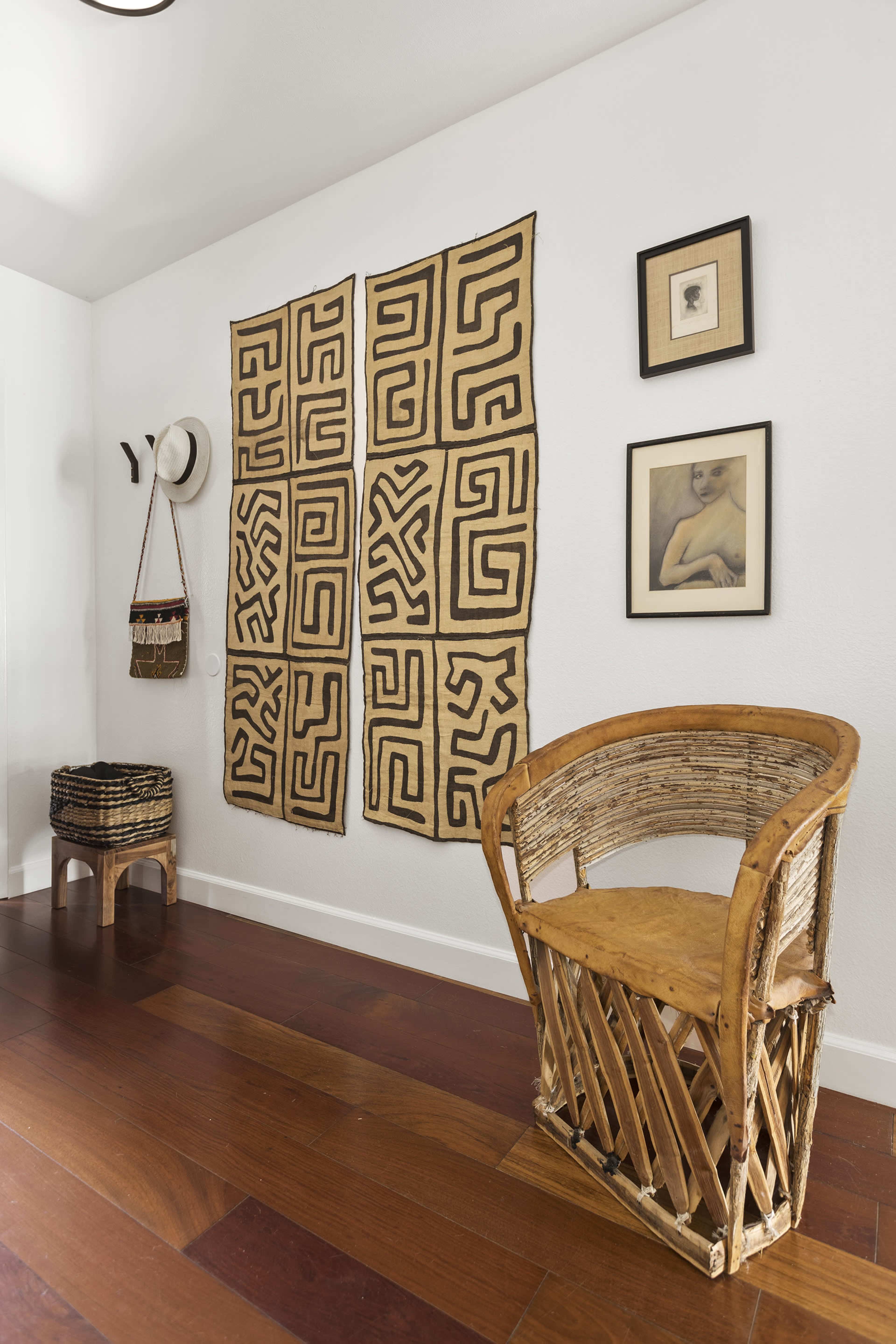 A corner of a room features a patterned wall hanging, a wooden chair, and framed artwork, with a hat and a bag hanging nearby.