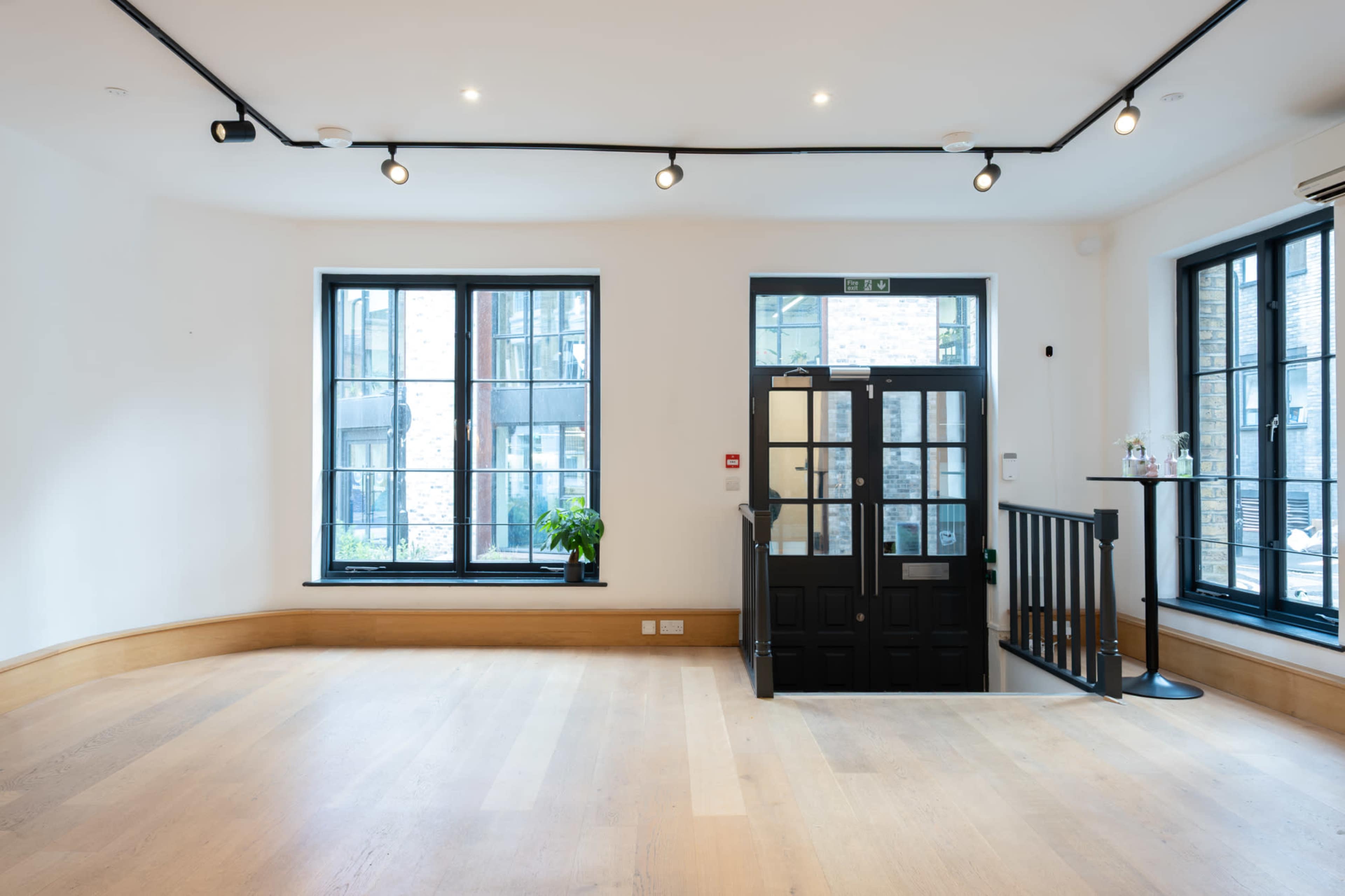 The image shows a spacious, well-lit room with large windows and a black door leading outside.
