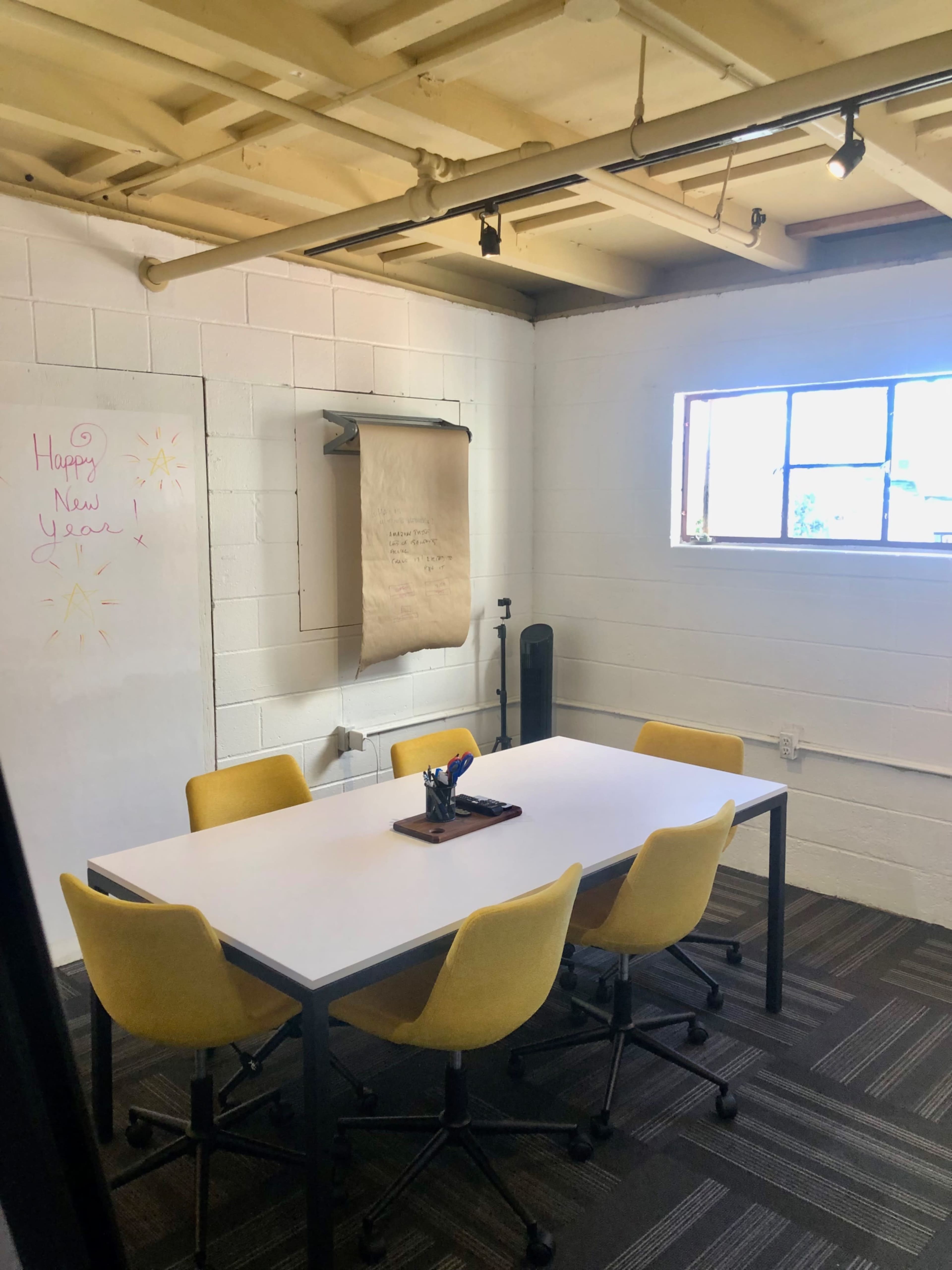 A conference room features a white table surrounded by yellow chairs, with a window on one wall and a large paper roll mounted for notes.