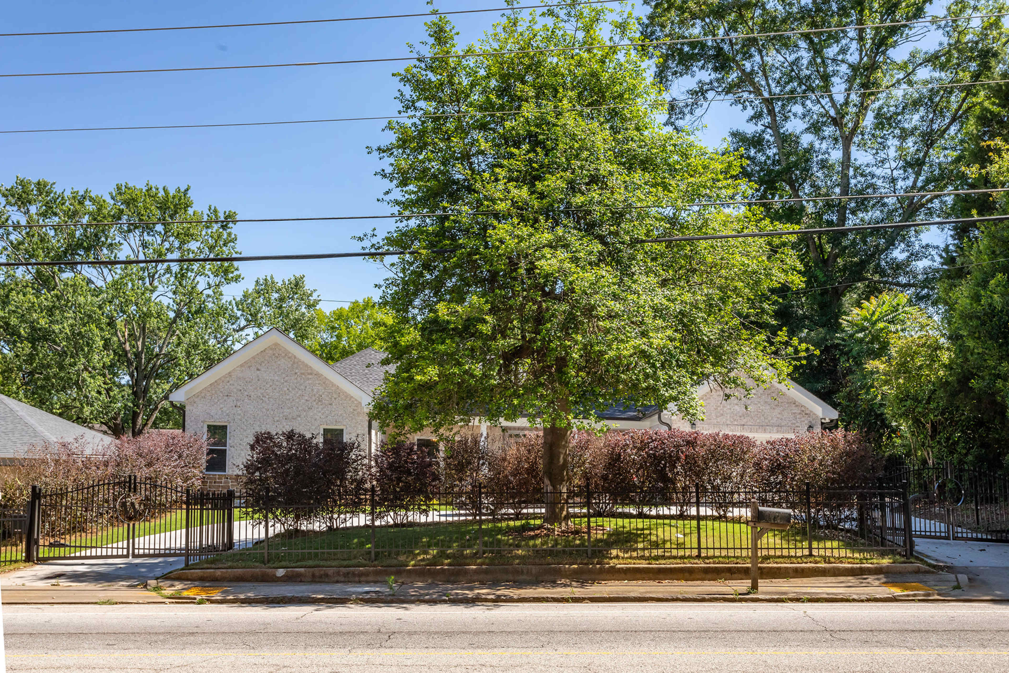 A single-story house with a landscaped yard is set behind a black wrought-iron fence and surrounded by various trees and shrubs.