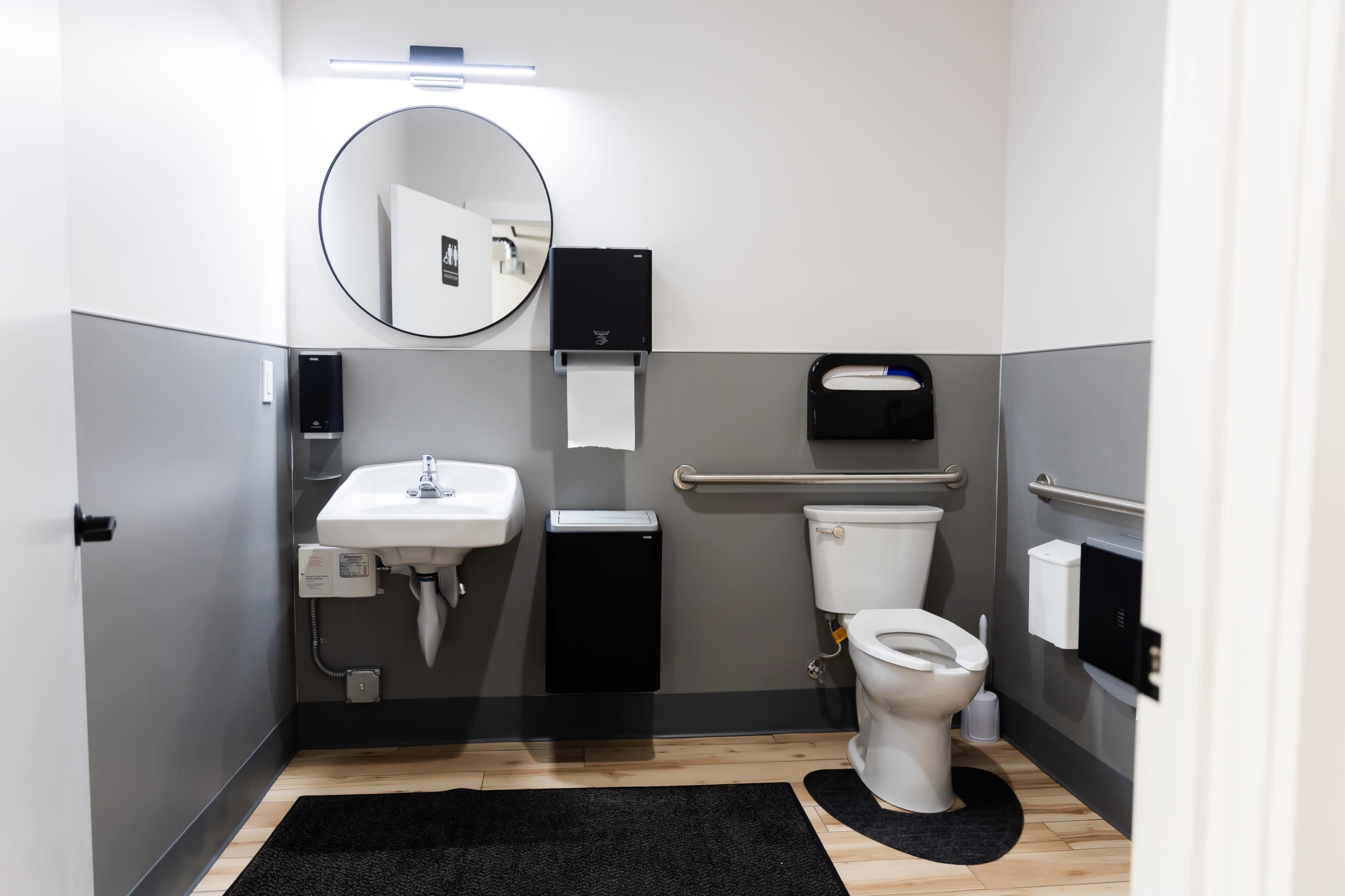 The image shows a modern restroom with a toilet, sink, mirror, and hand dryer, set against a two-tone gray wall and wooden floor.