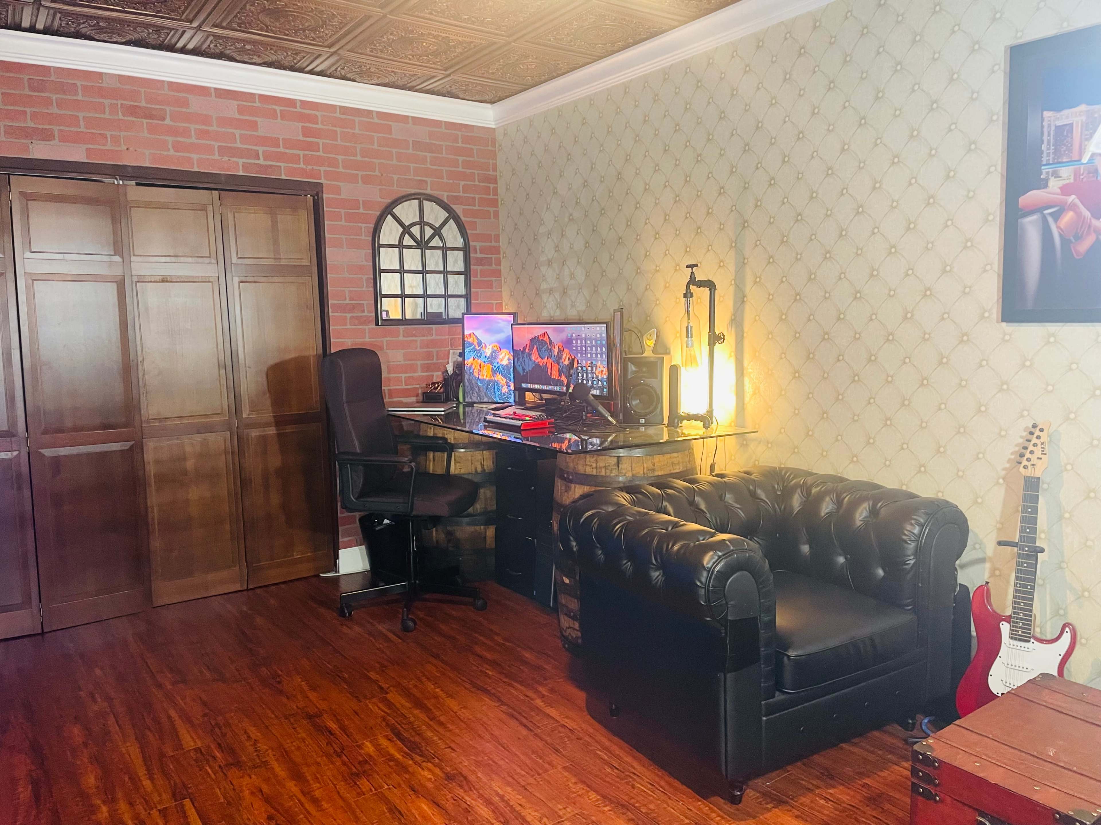 The image shows a home office with a black leather chair, a glass desk featuring a computer monitor, and a guitar leaning against a wooden crate, all set against a patterned wallpaper and a brick wall.