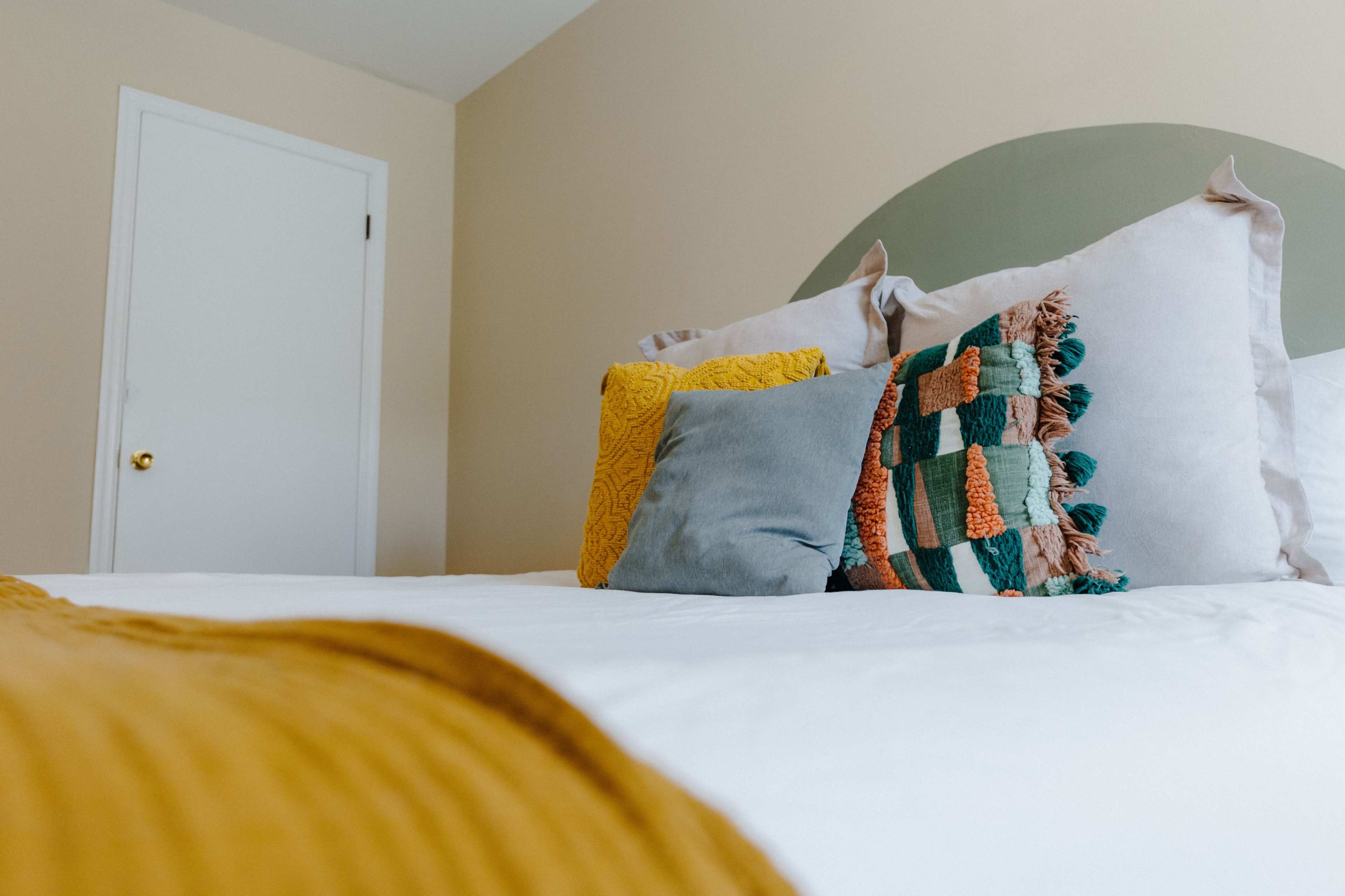 A neatly made bed with decorative pillows in shades of yellow and teal is positioned in a softly lit bedroom with a closed door in the background.