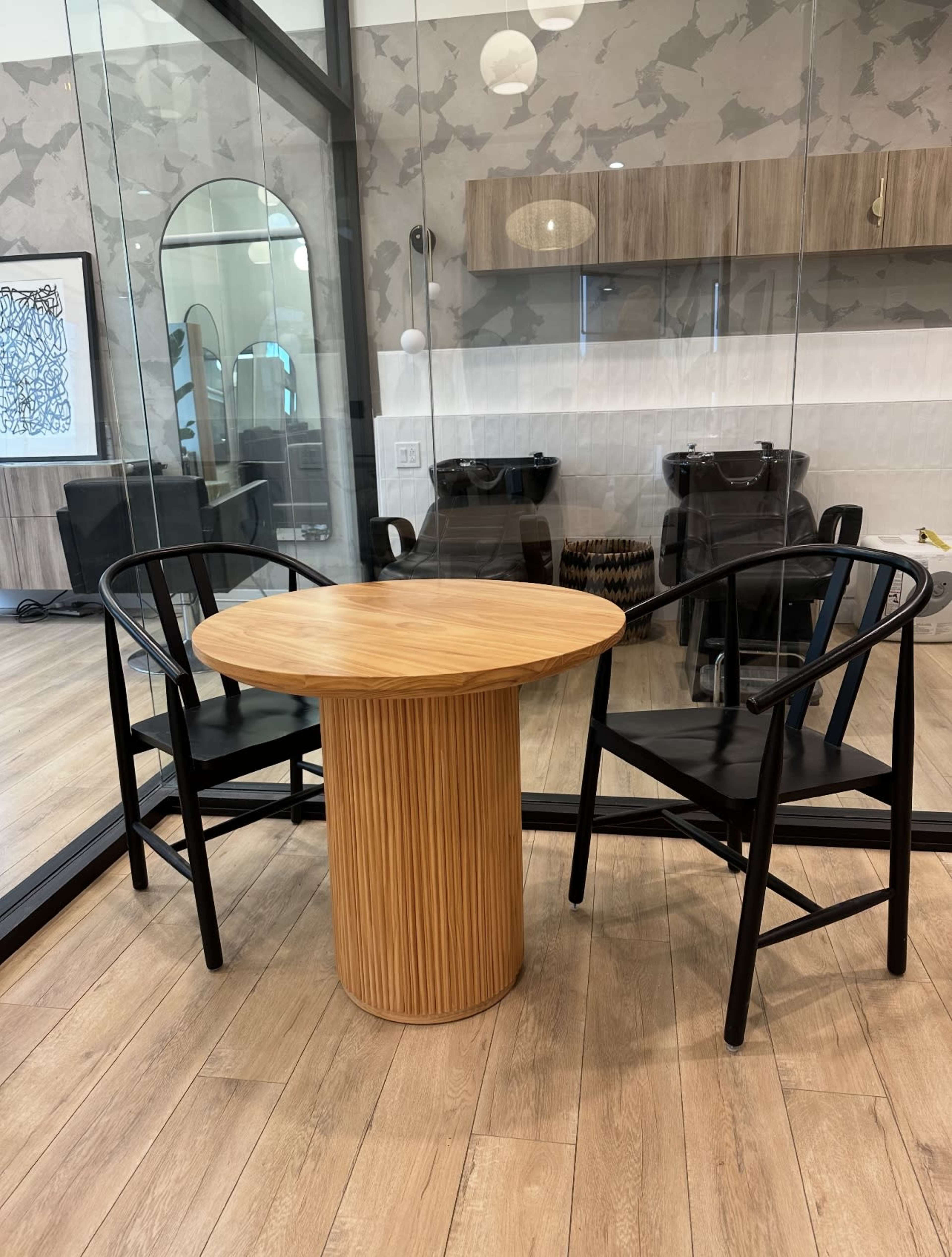 The image shows a round wooden table with vertical grooves and two black chairs in a modern, glass-walled interior space.