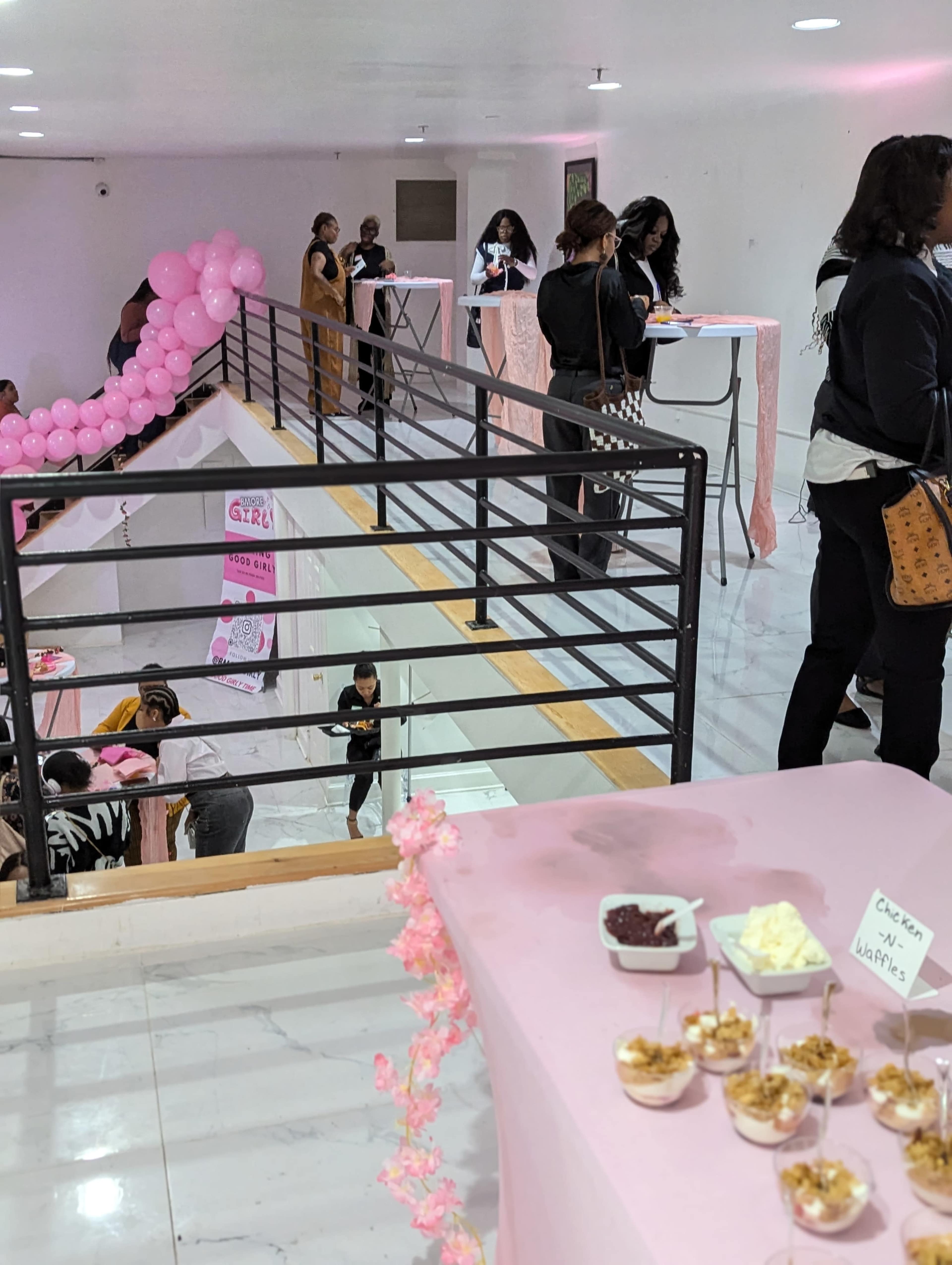 The image shows a spacious event venue decorated with pink balloons, where attendees engage at high tables on a balcony level while others gather below at a refreshment station.