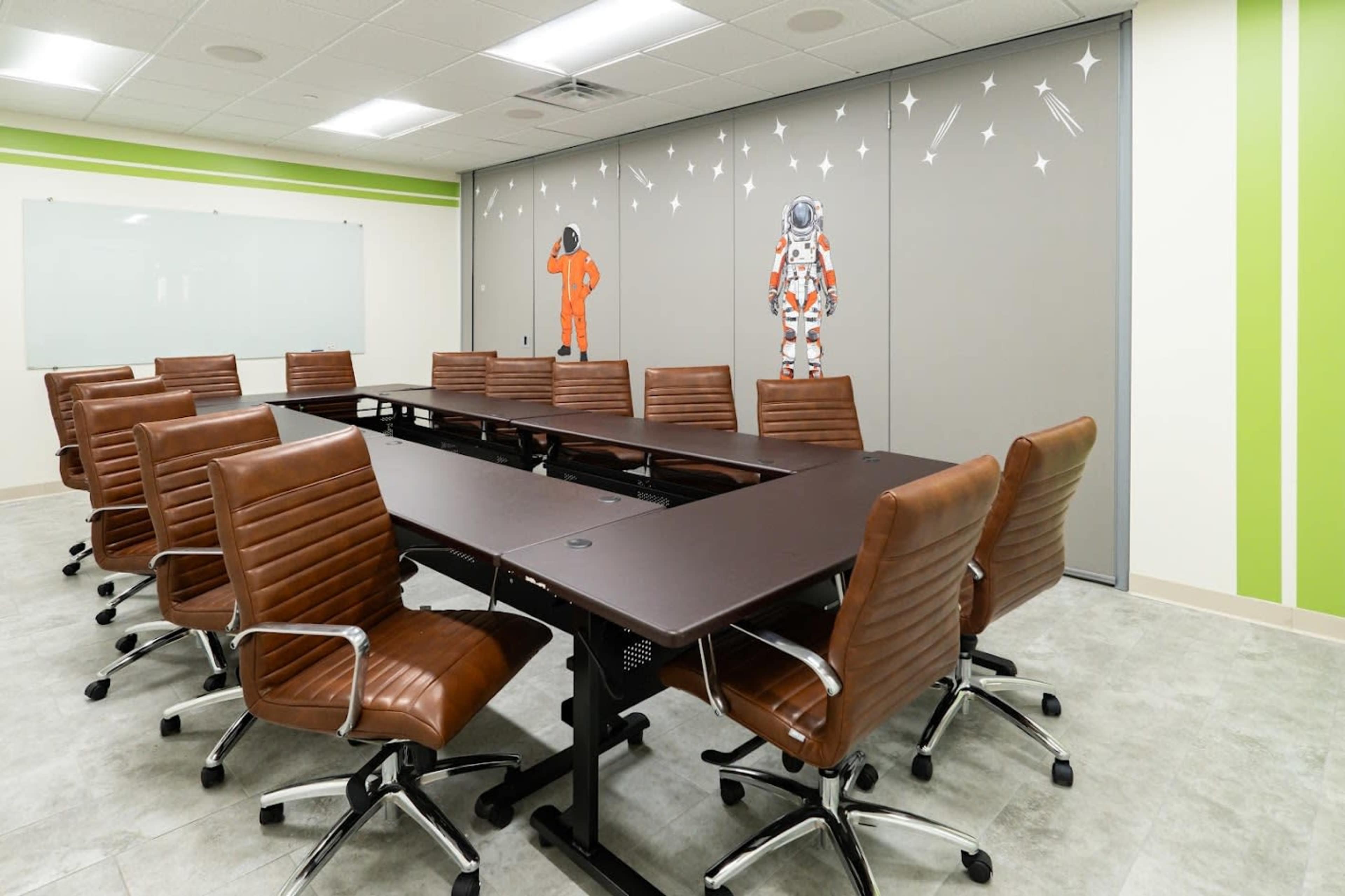 The image shows a modern conference room with a long dark table surrounded by brown leather chairs and wall art of astronauts.