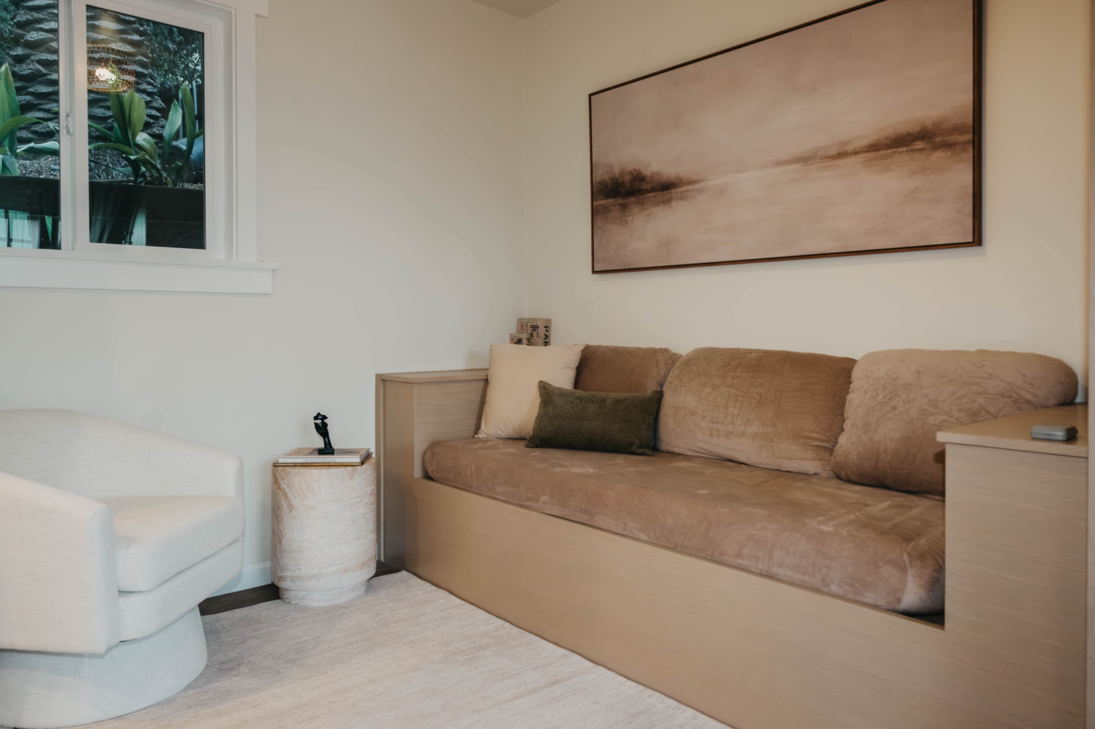 The image shows a cozy corner of a room with a beige couch, a light-colored chair, and a large artwork hanging on the wall.