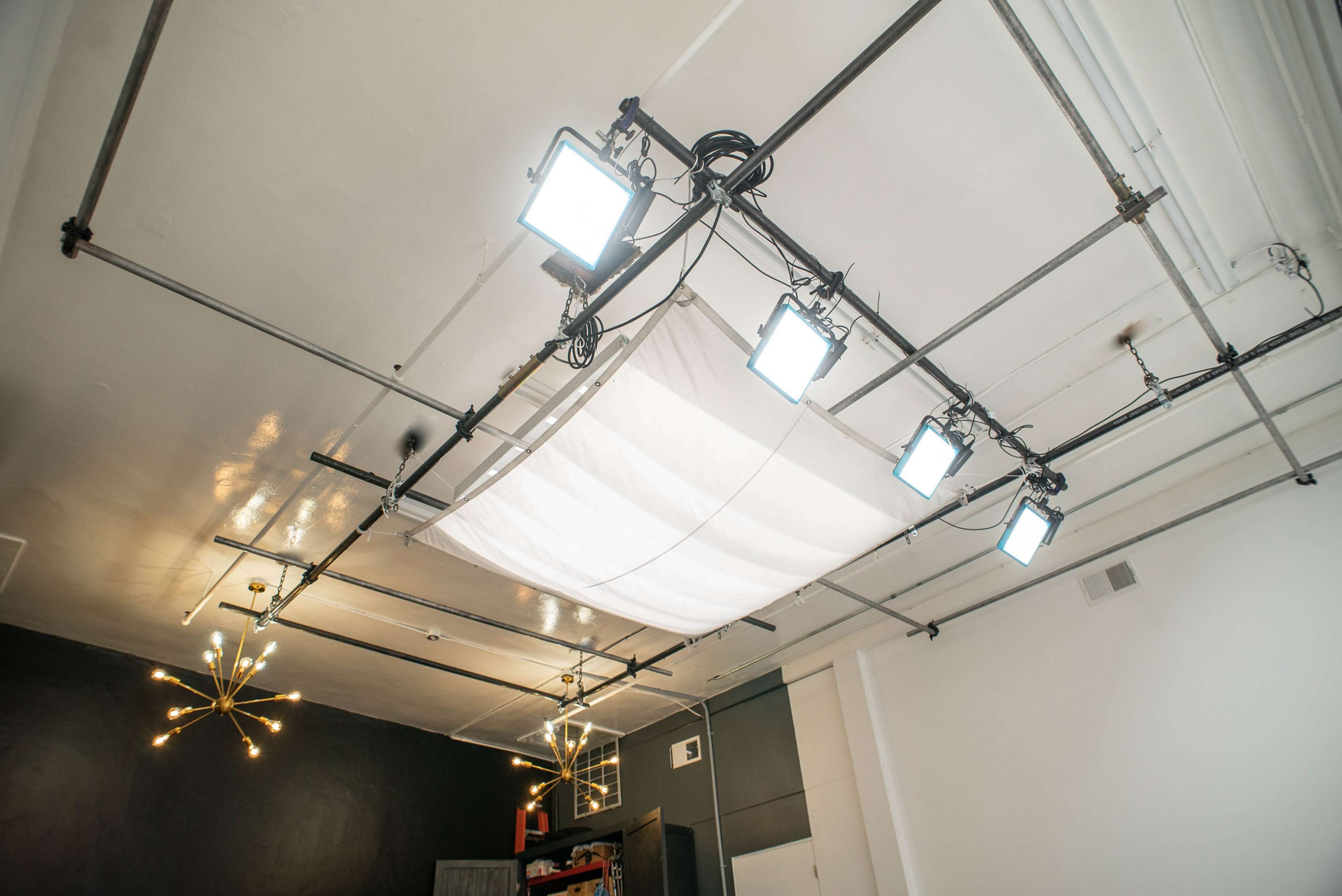A ceiling is fitted with multiple lights and a large fabric diffuser, surrounded by industrial-style metal piping and fixtures.