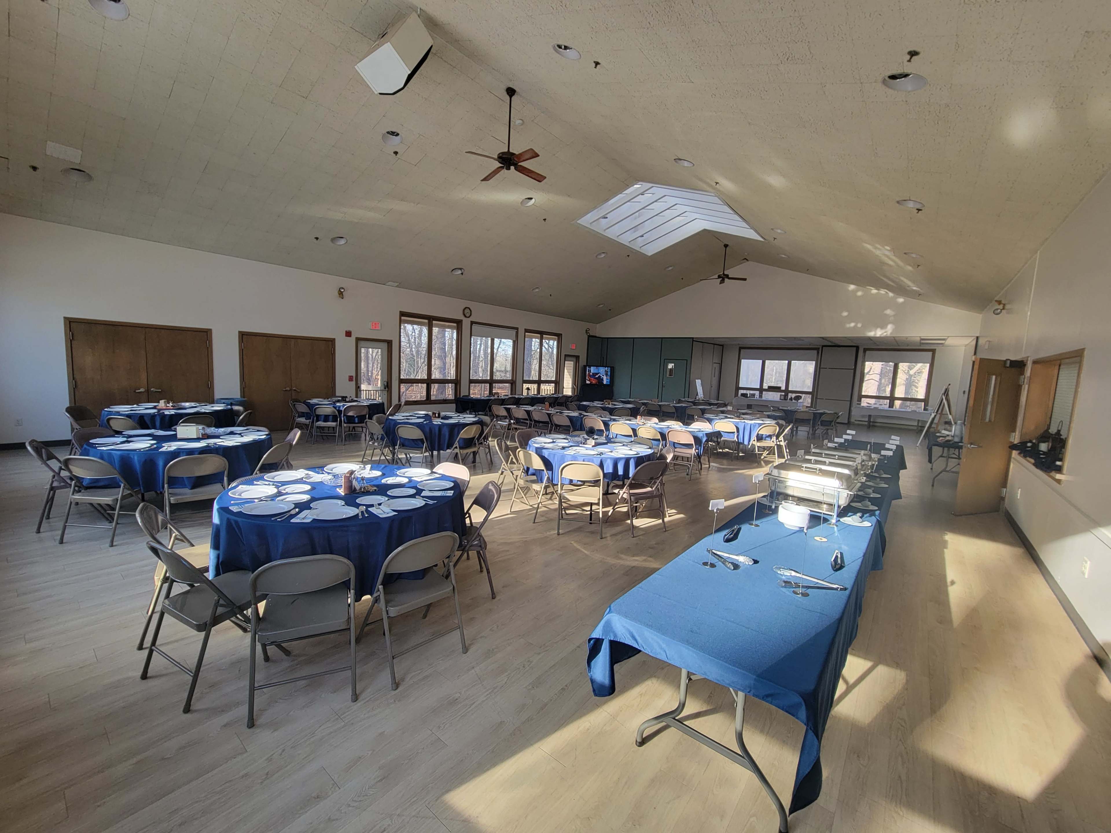 The image shows a spacious banquet hall set up with round tables covered in blue tablecloths, a buffet area, and large windows allowing natural light to enter.