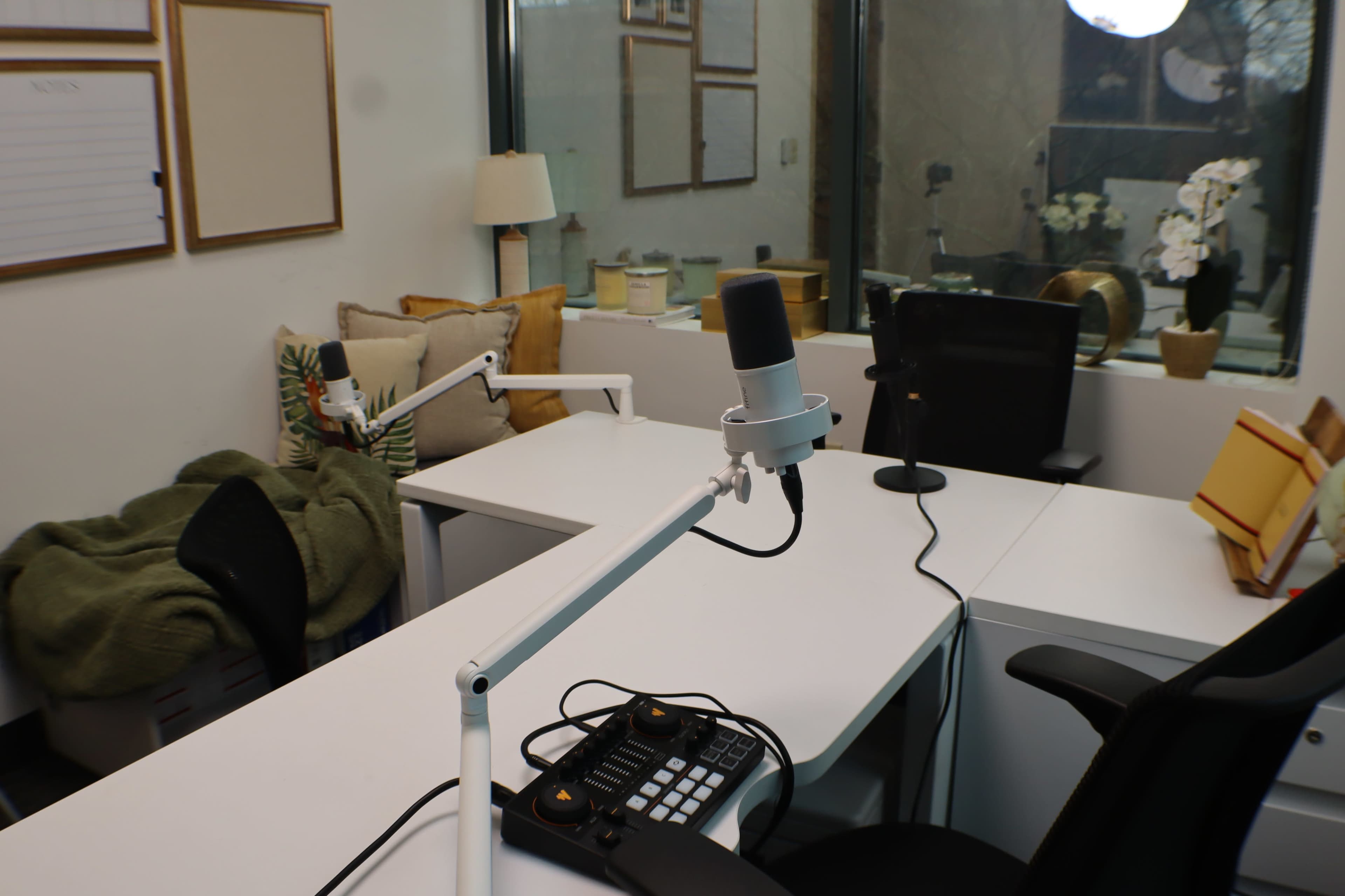 The image shows a modern recording studio with two desks, a microphone setup, and a cozy seating area featuring cushions and a lamp.