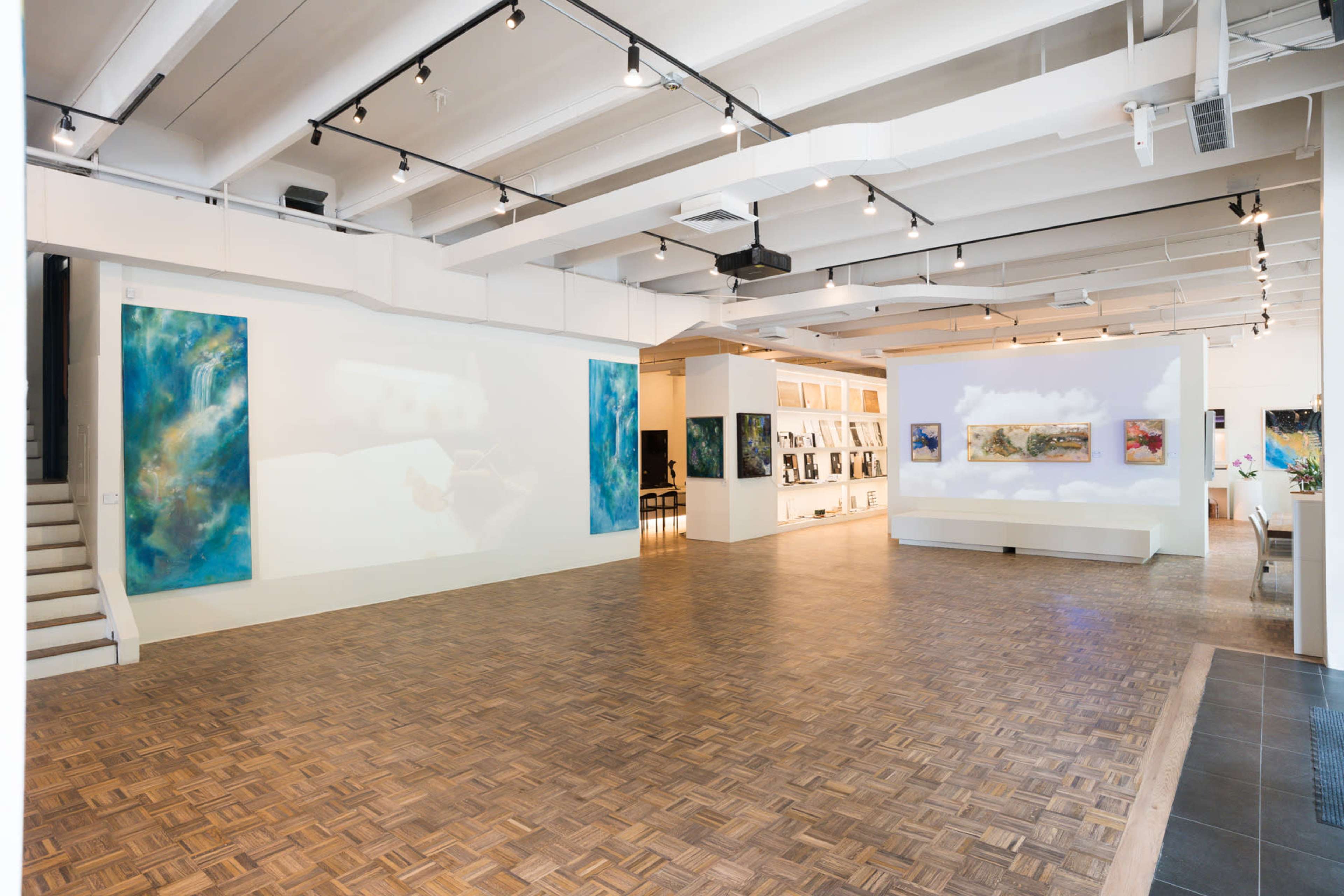 The image shows a spacious art gallery featuring abstract paintings on the walls, a wooden floor, and a white seating area against a backdrop of projected images.