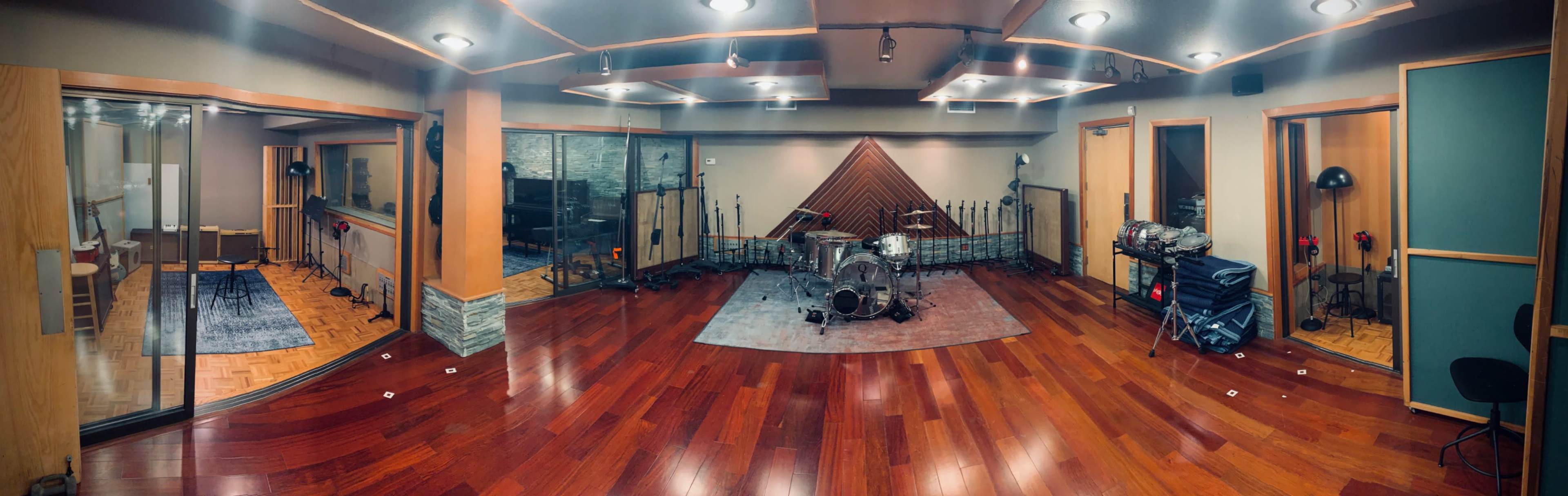 A panoramic view of a professional recording studio featuring a drum set on a rug and soundproof booths along the walls.