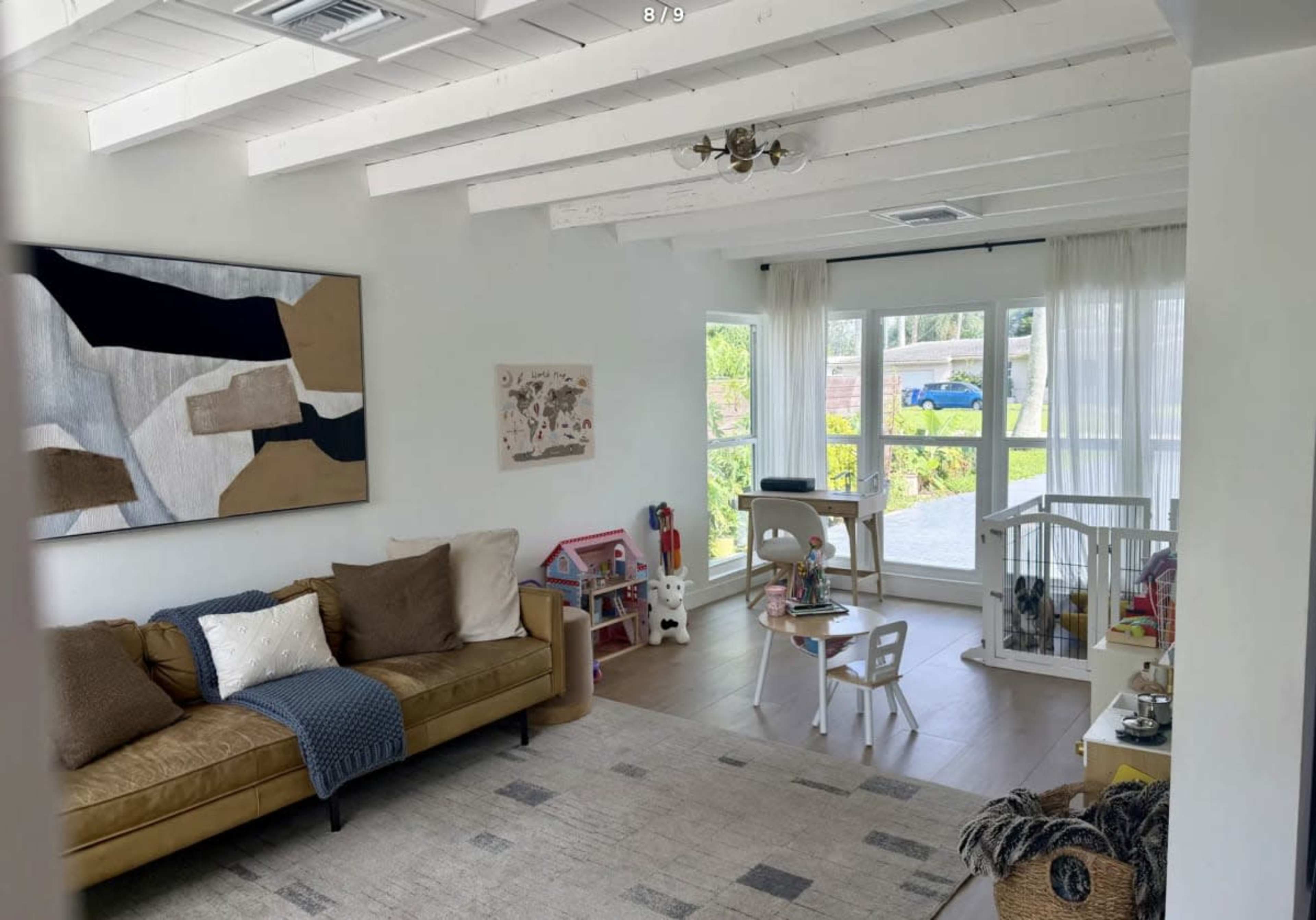 The image shows a bright living room with a brown sofa, a patterned wall art piece, and children's toys scattered around, along with a window that provides natural light.