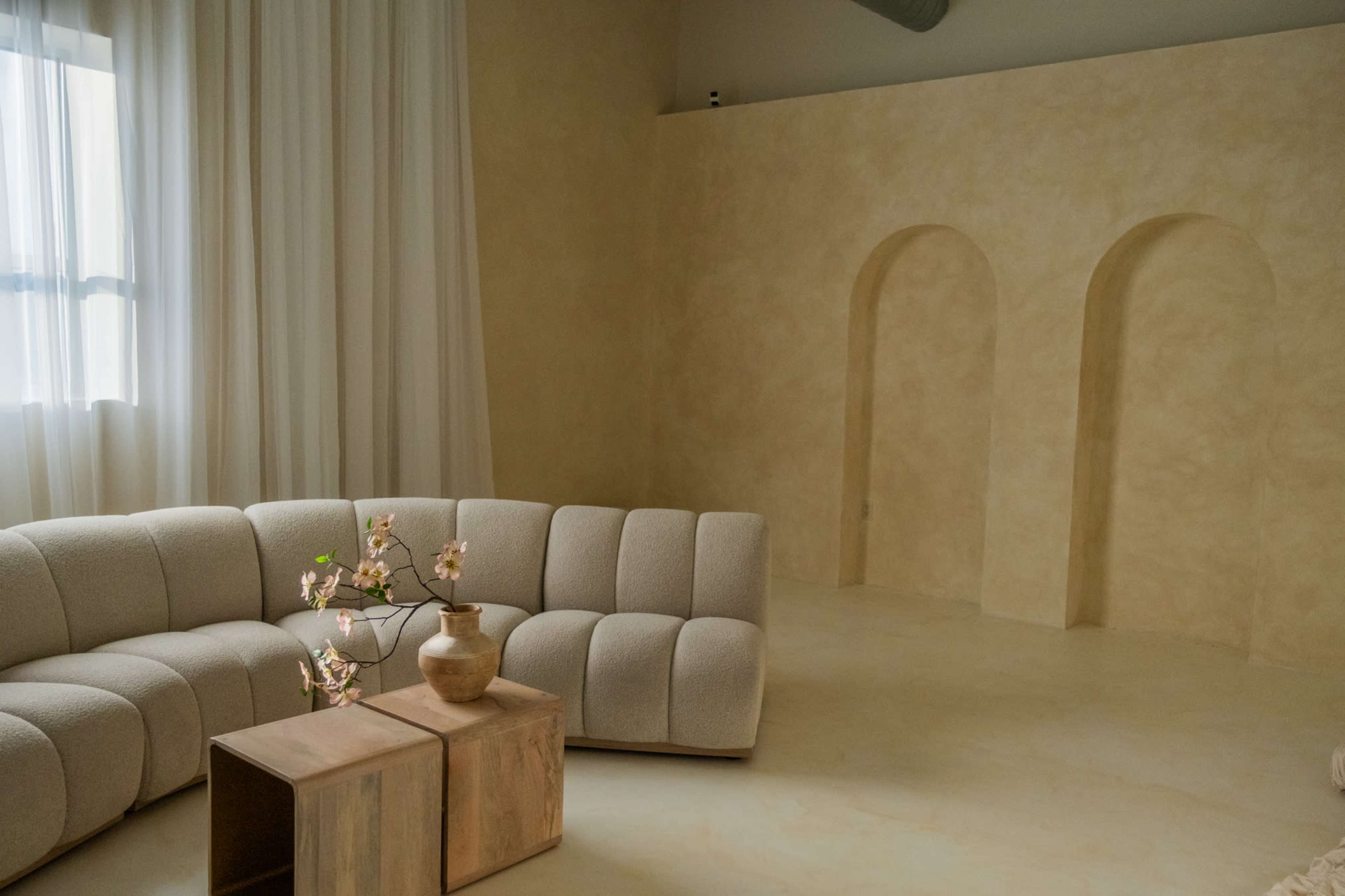The image shows a minimalist living room featuring a curved sofa, two wooden side tables, and an arched architectural feature against a neutral-toned wall.