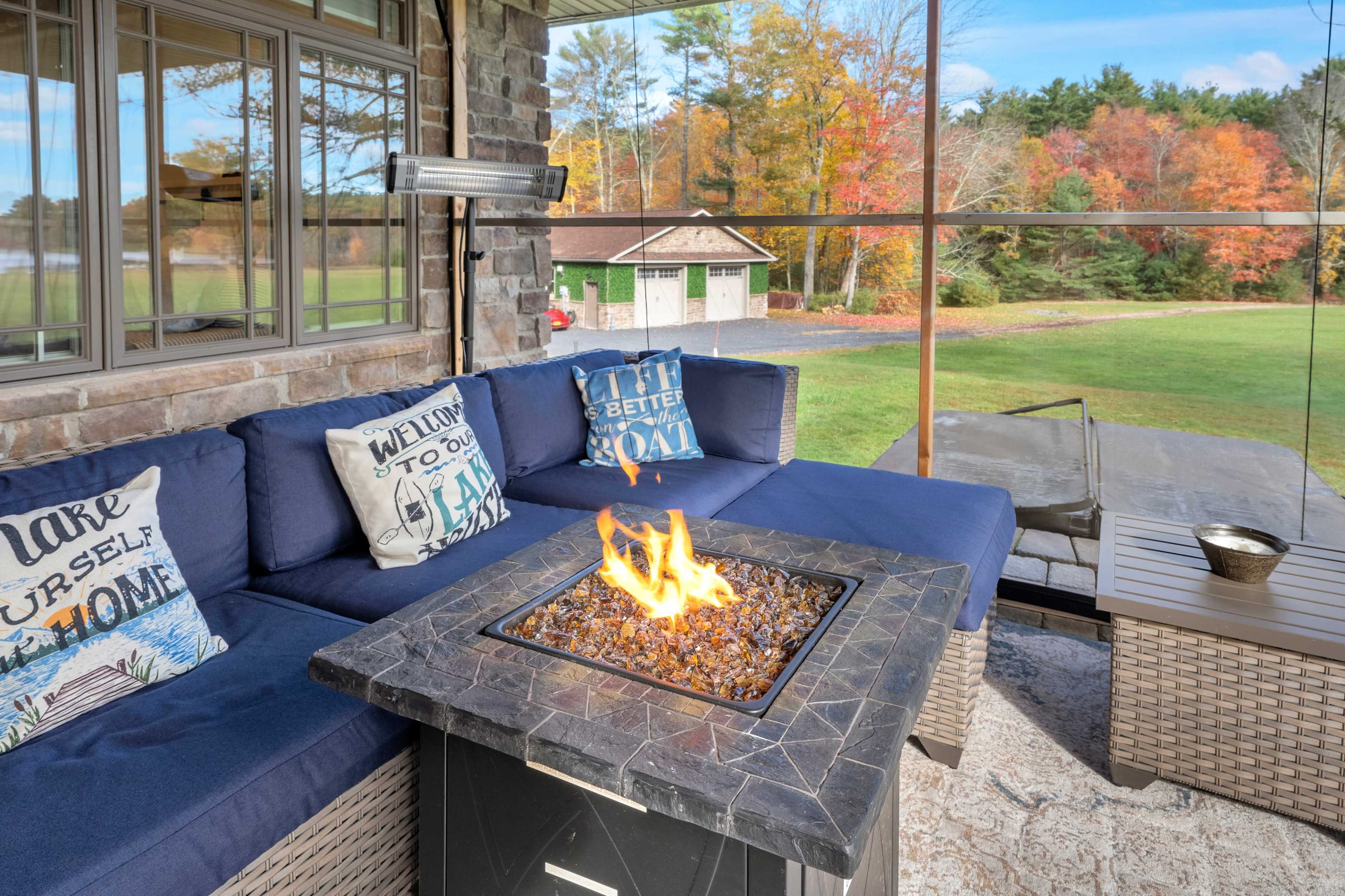 A cozy outdoor seating area features a fire pit surrounded by blue cushions, with a view of a grassy yard and trees in autumn colors.