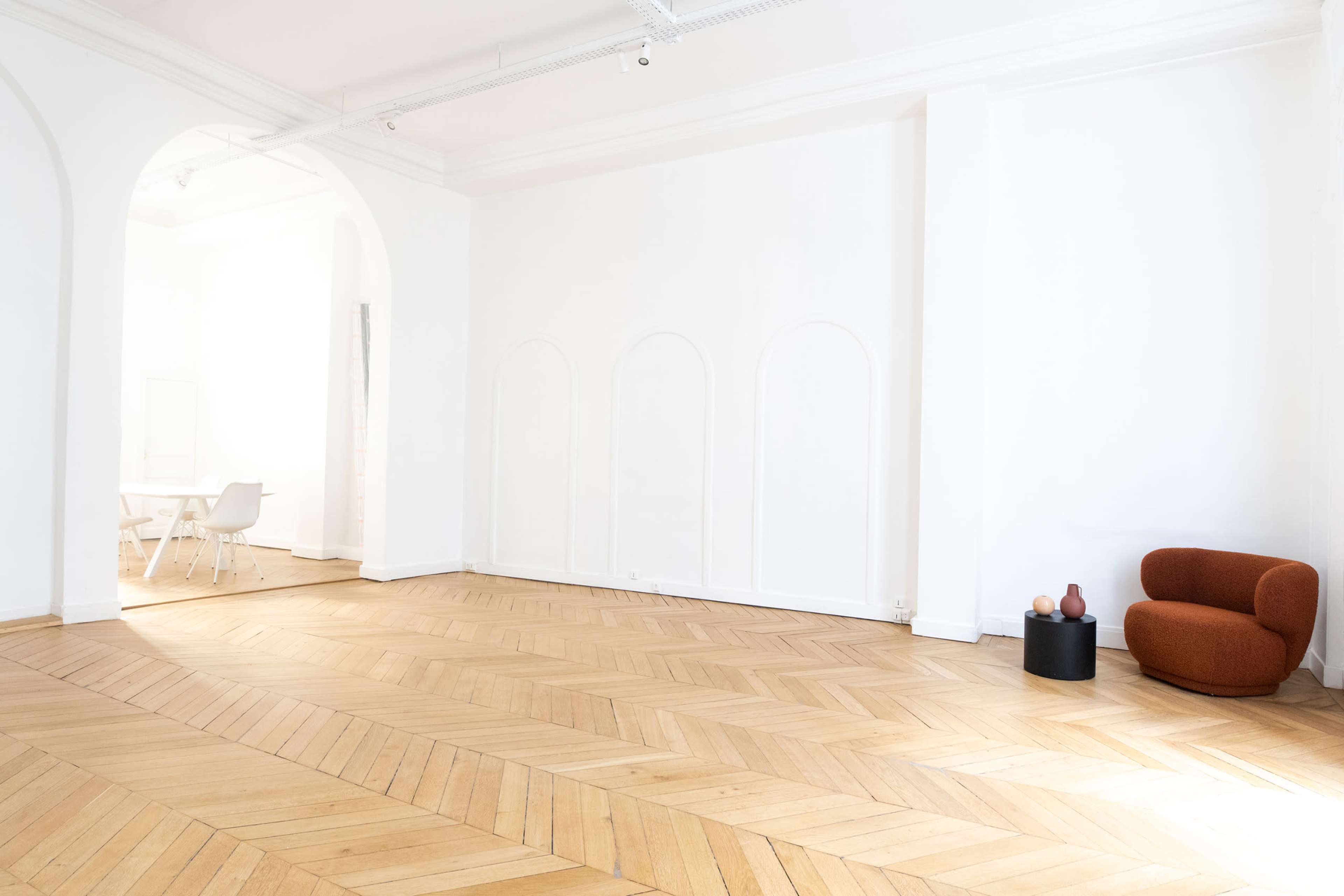 A spacious, minimally furnished room with wooden herringbone flooring, featuring an orange armchair and a small black table in one corner.