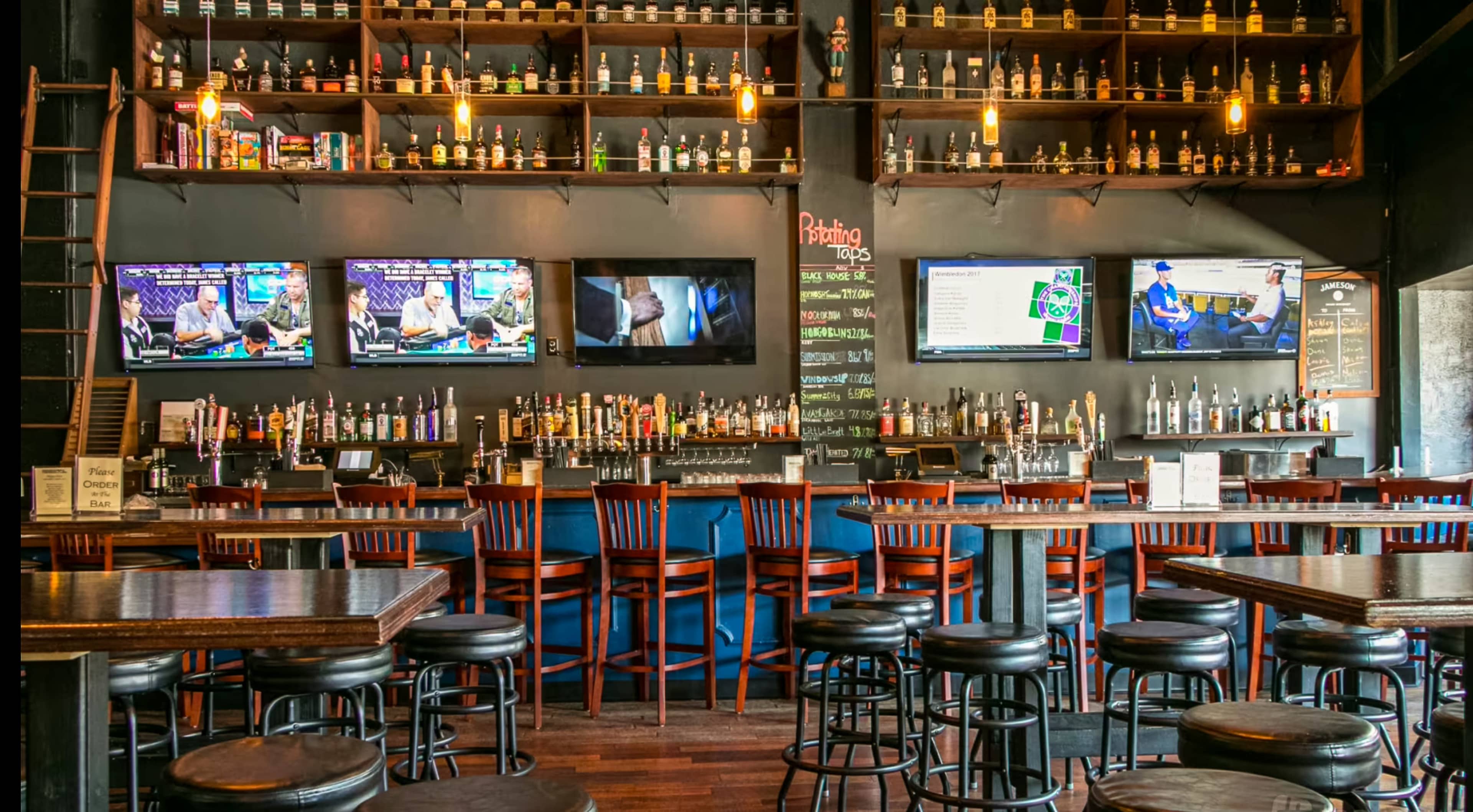 The image shows a bar with rows of bottles on shelves, several televisions displaying sports, and wooden tables and chairs arranged around the space.