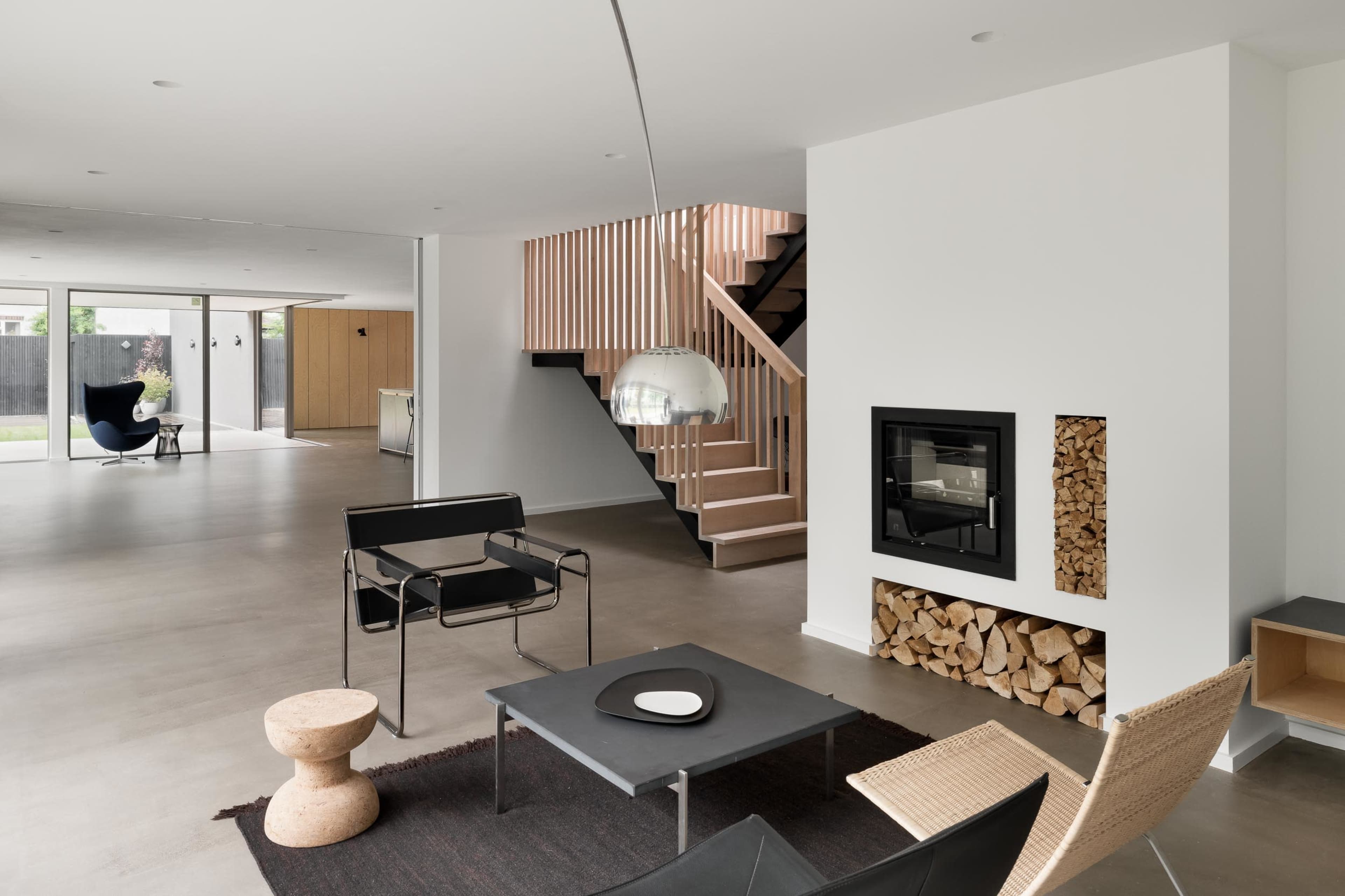 A modern living space featuring a minimalist design with a sitting area that includes two chairs, a table, and a fireplace surrounded by stacked wood.