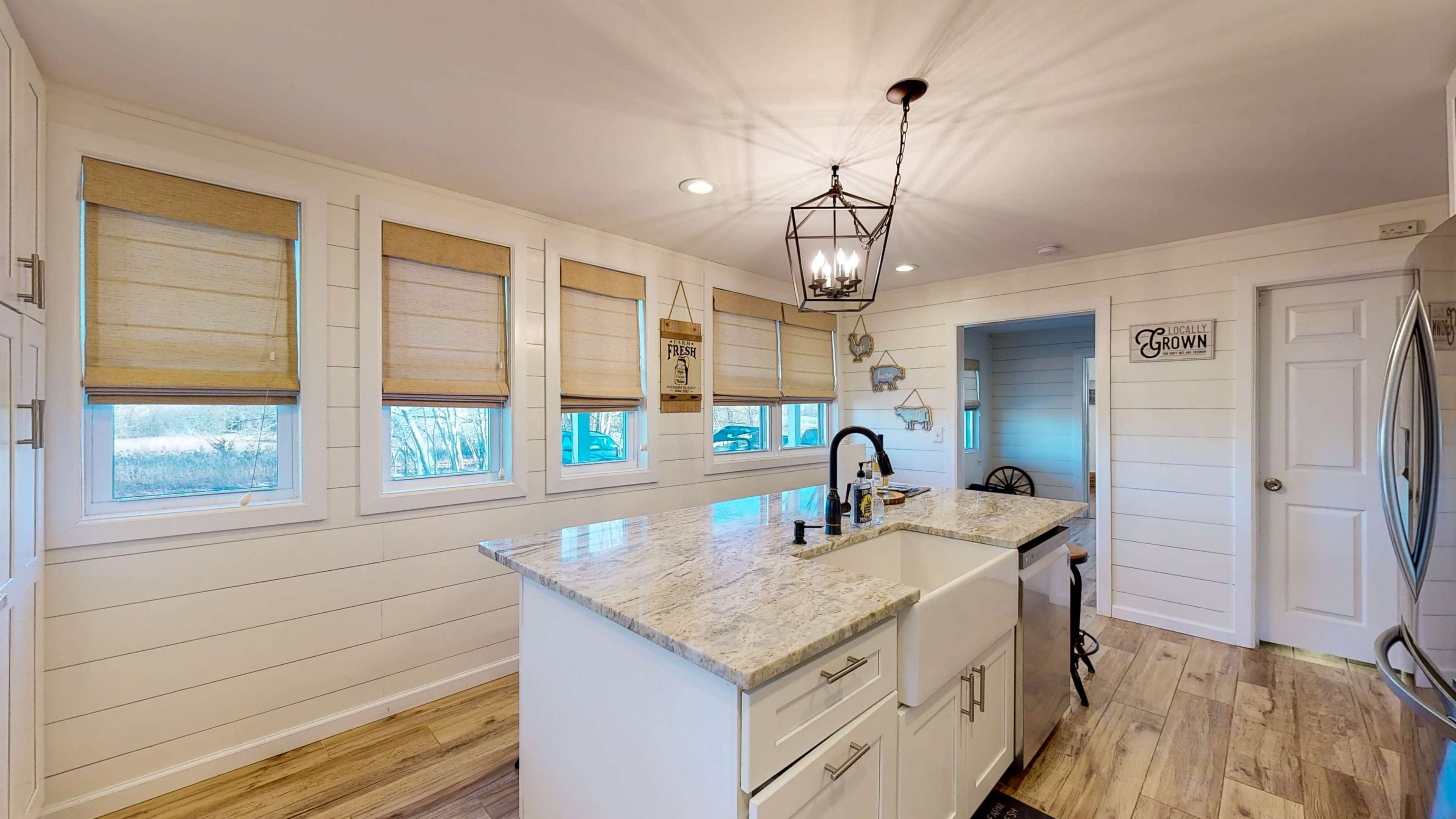 The kitchen features a central island with a granite countertop, white cabinetry, and multiple windows adorned with Roman shades.