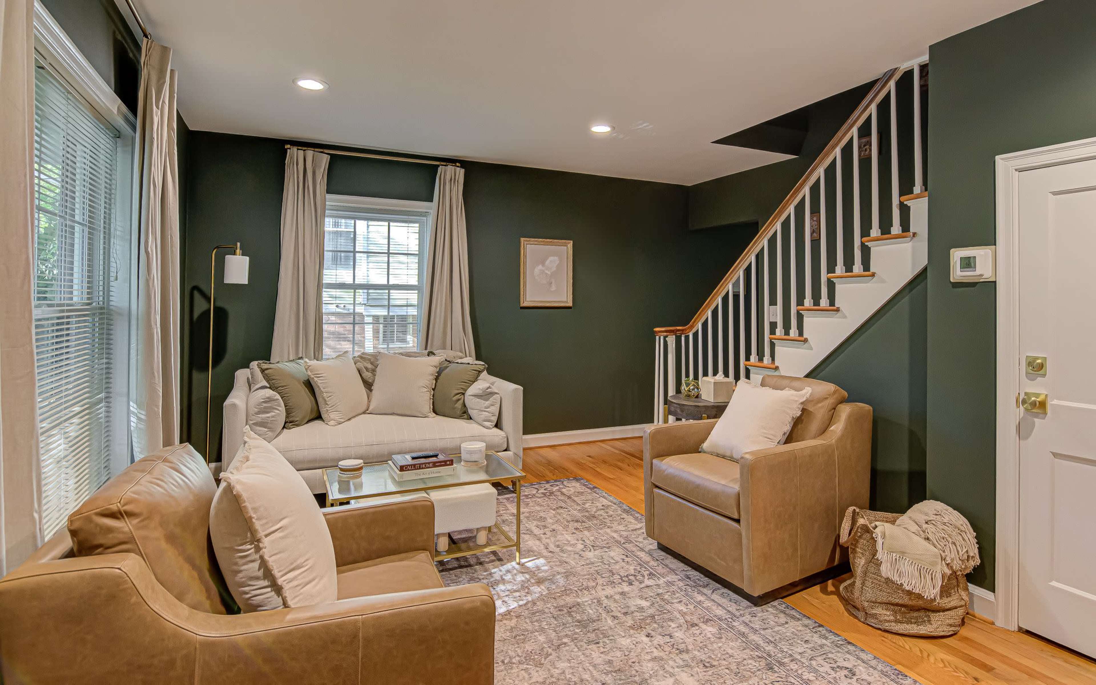 The image shows a living room with two leather armchairs, a light-colored sofa, a coffee table, and a staircase in a home with dark green walls.