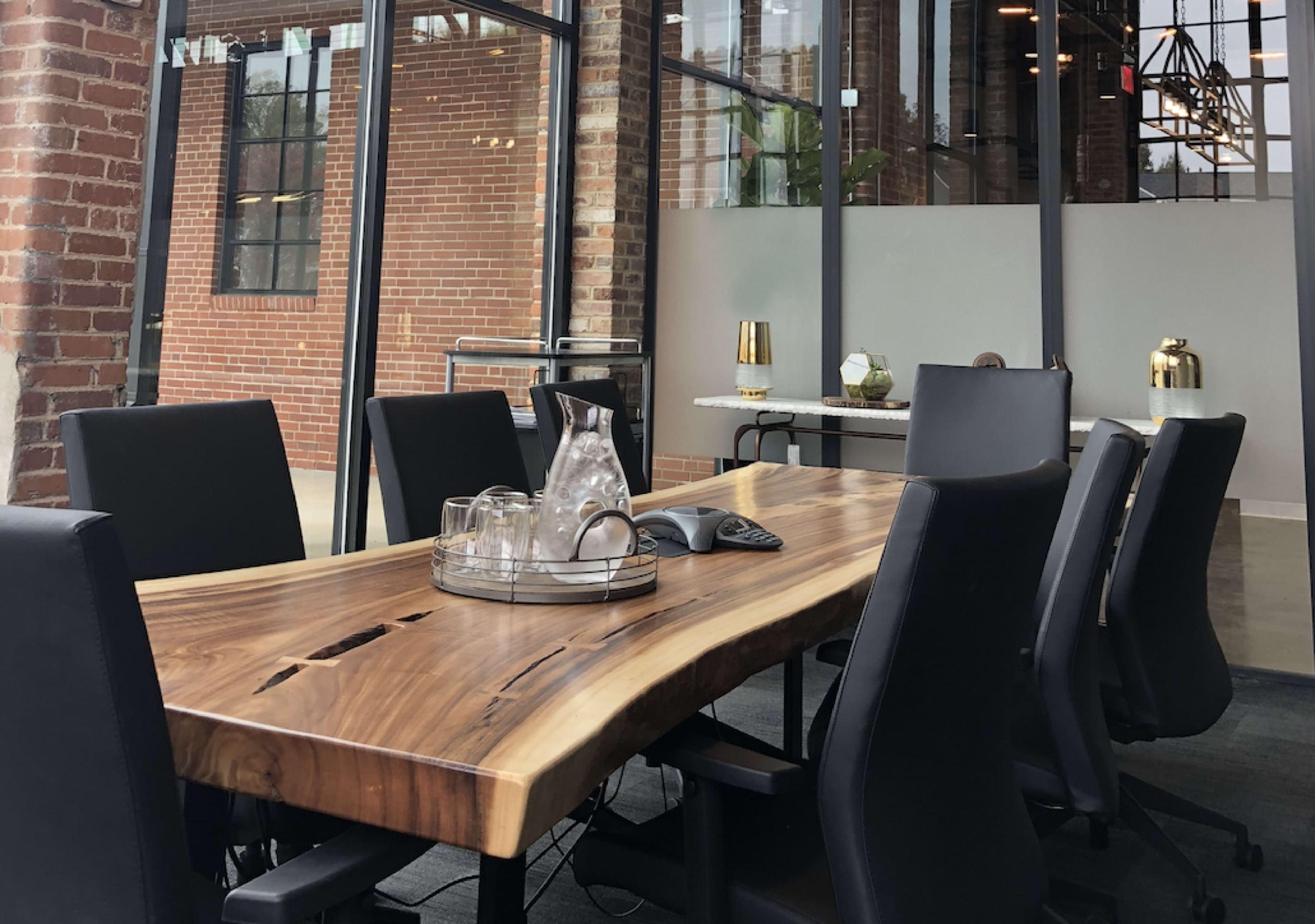 A wooden conference table with black chairs is set up in a spacious meeting room featuring exposed brick walls and large windows.