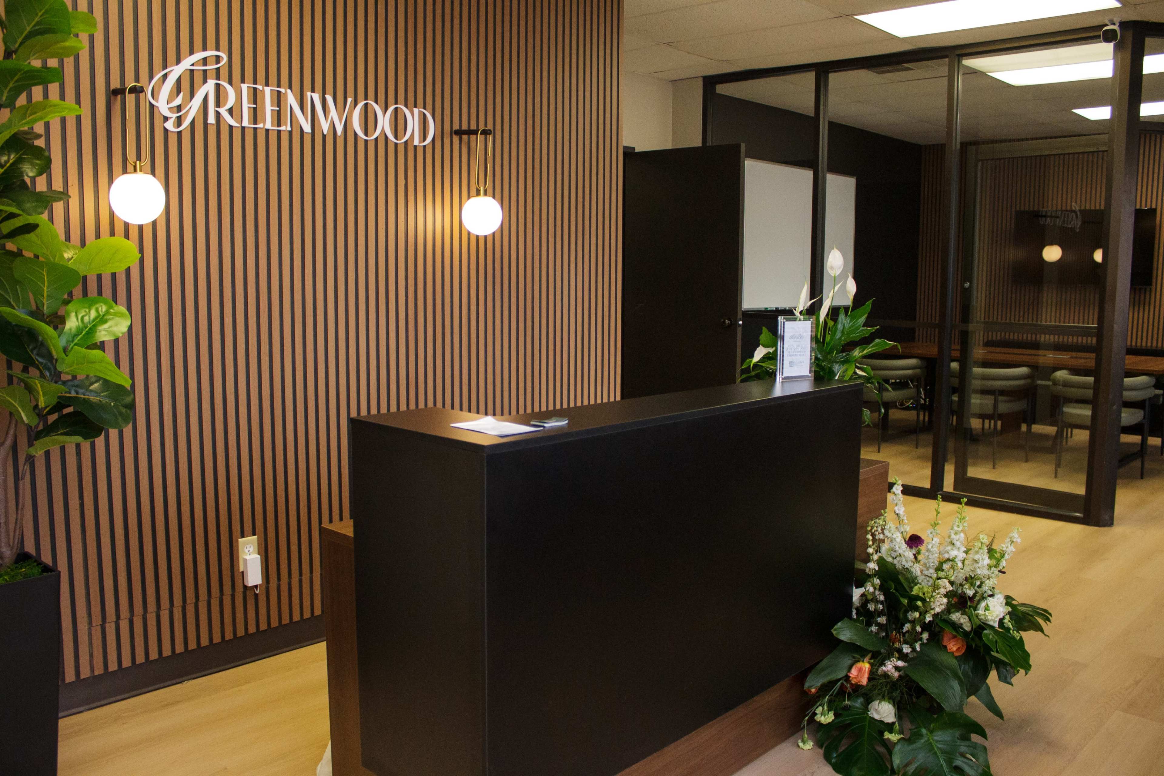 The image shows a modern reception area featuring a dark wooden reception desk, a wall with vertical wooden slats, and a sign that reads "GREENWOOD."