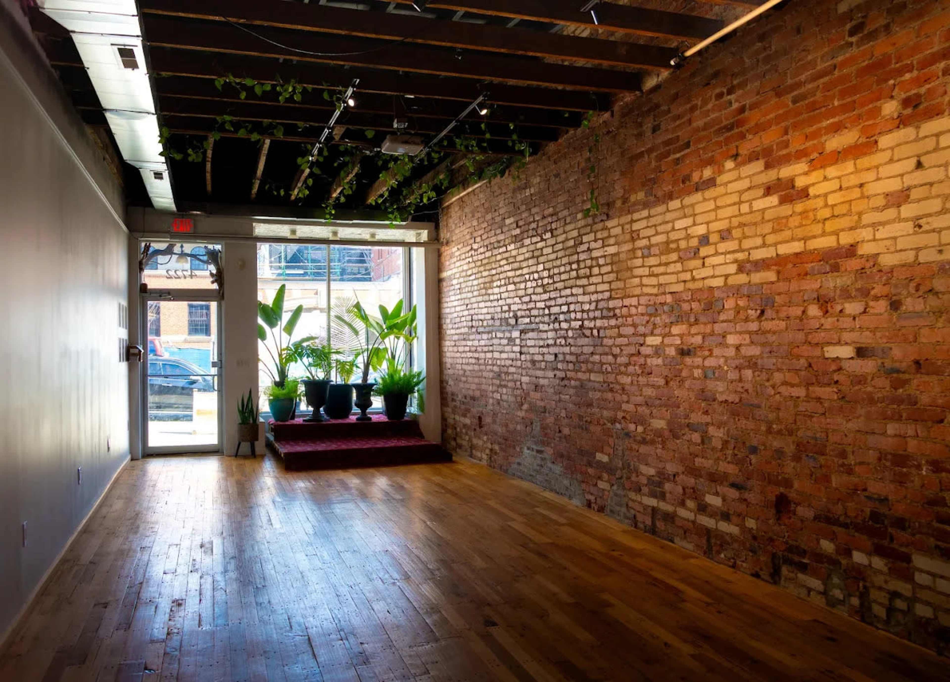 The image shows an empty room with exposed brick walls, wooden floors, and potted plants near a large window at the entrance.