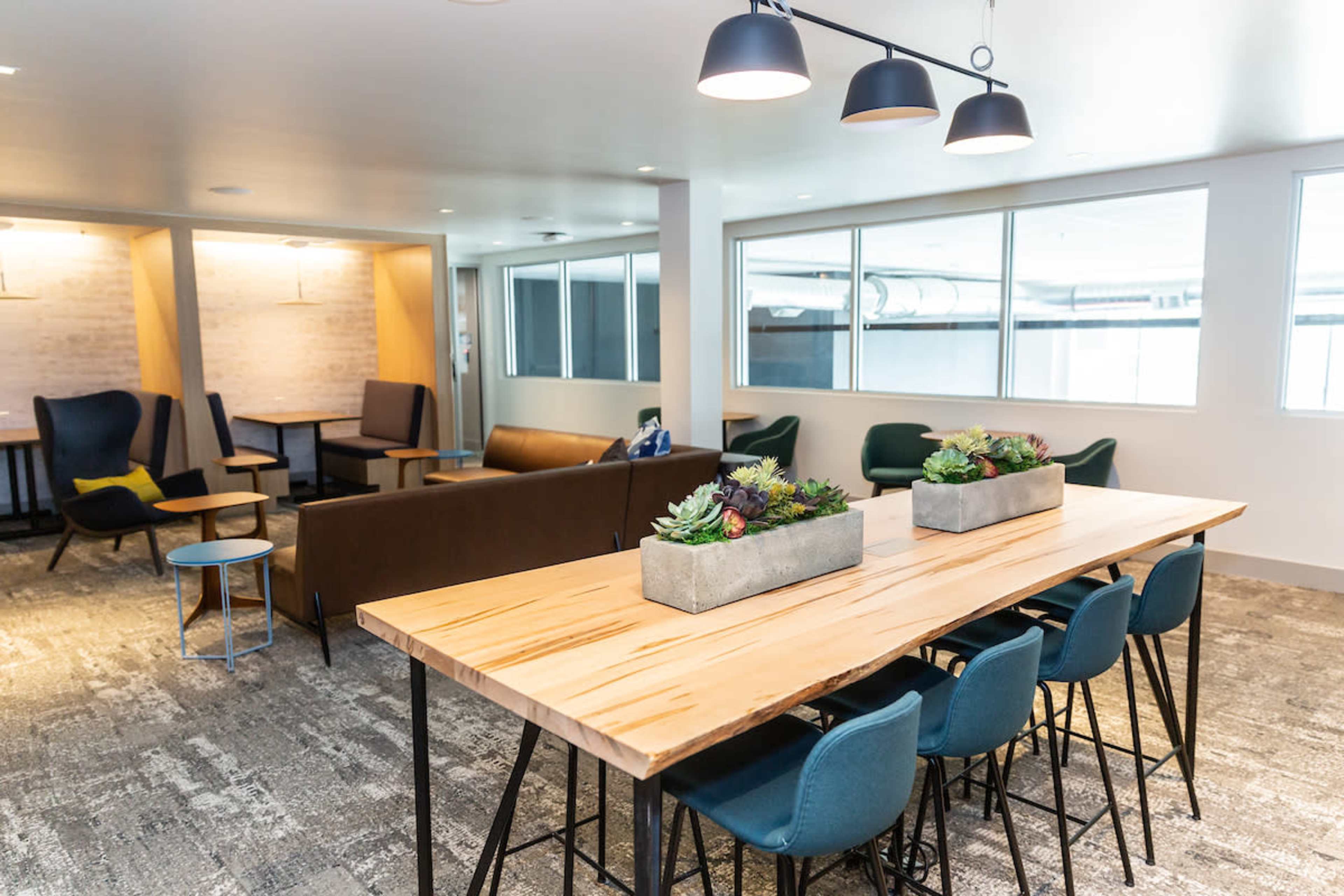 A modern lounge area features a long wooden table surrounded by chairs, with a seating arrangement of couches and chairs in the background.