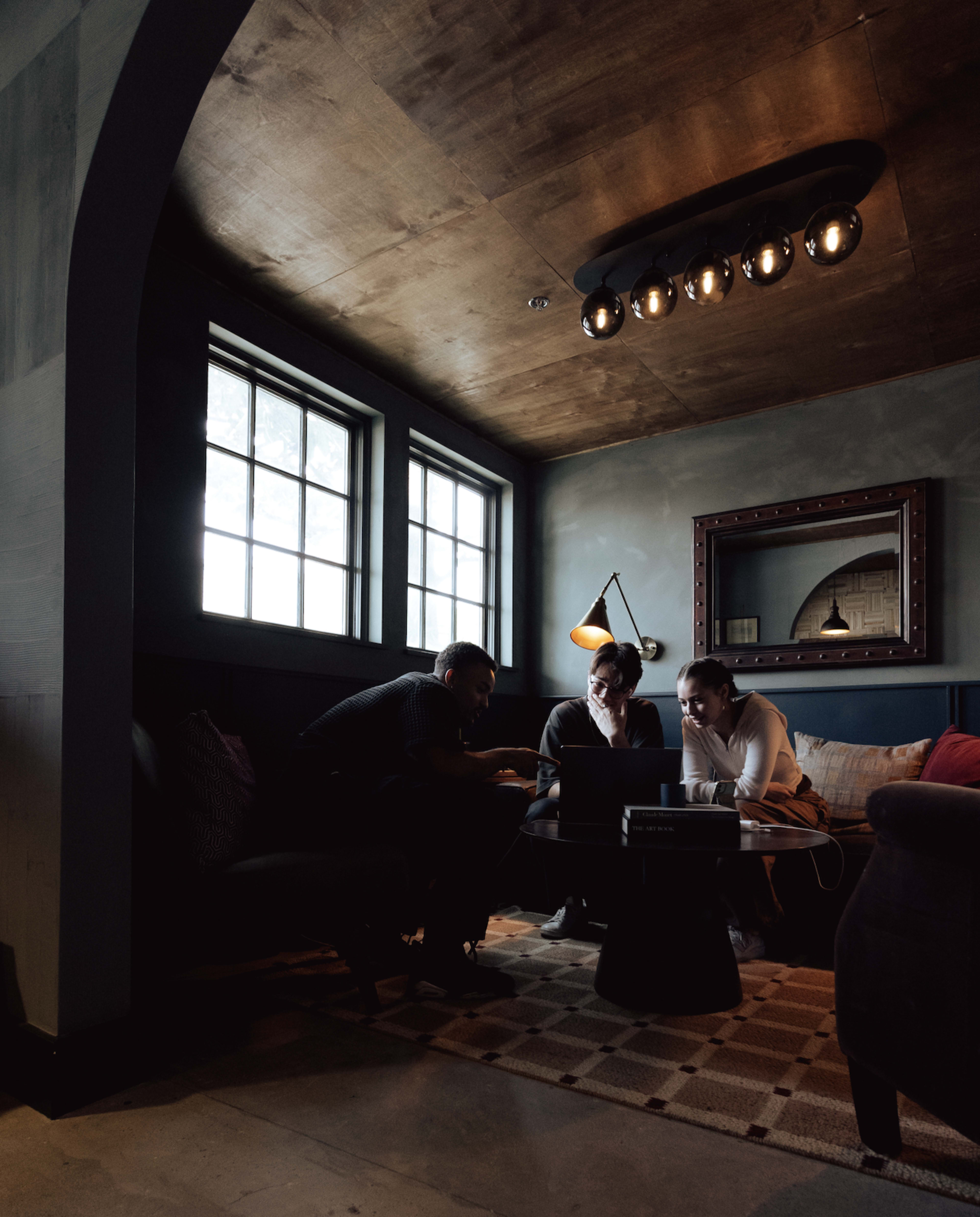Three people are engaged in discussion while seated on a couch in a dimly lit room with large windows and wooden accents.