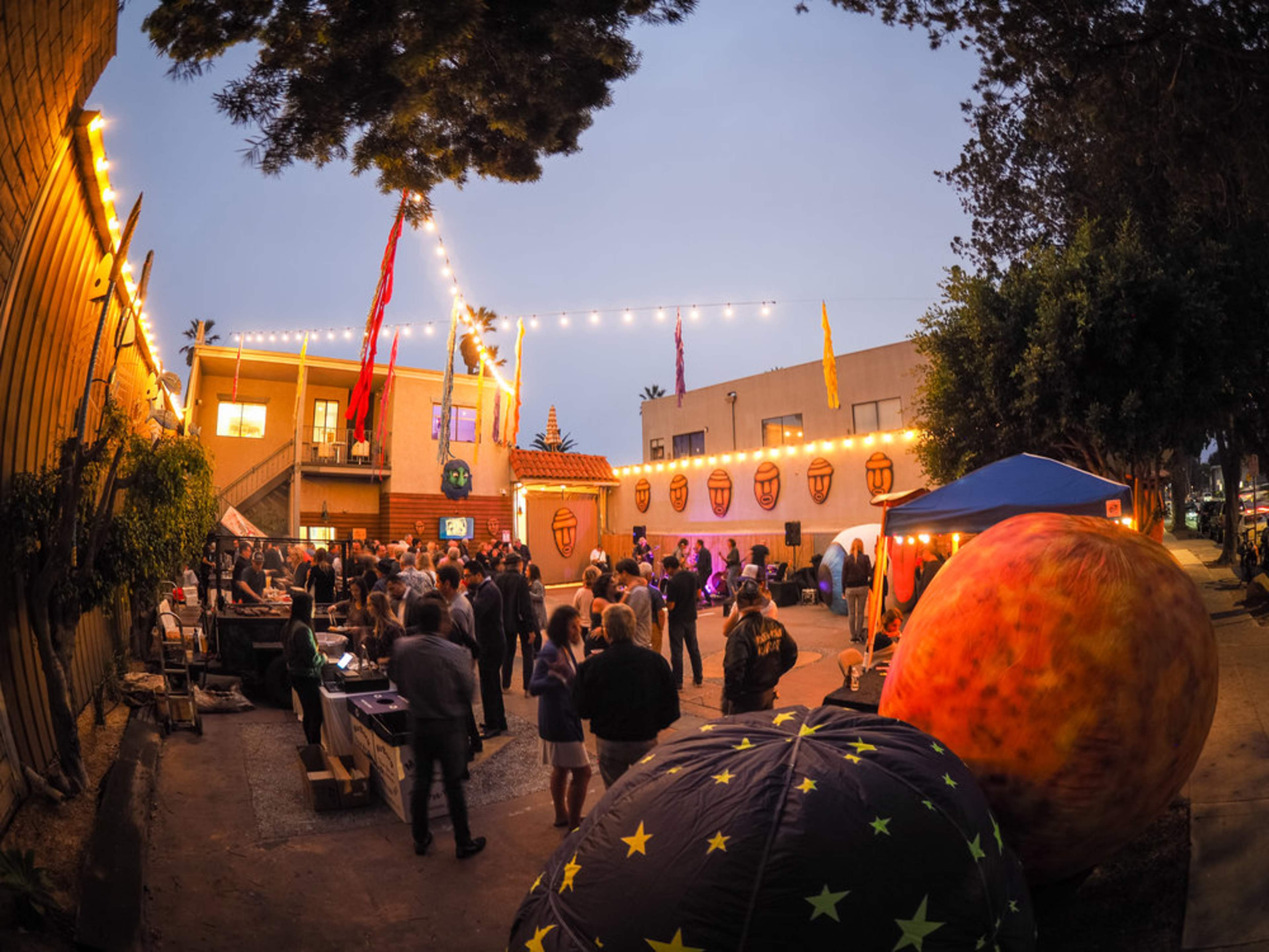 A crowd mingles in an outdoor space decorated with lights and banners during a social event in the evening.