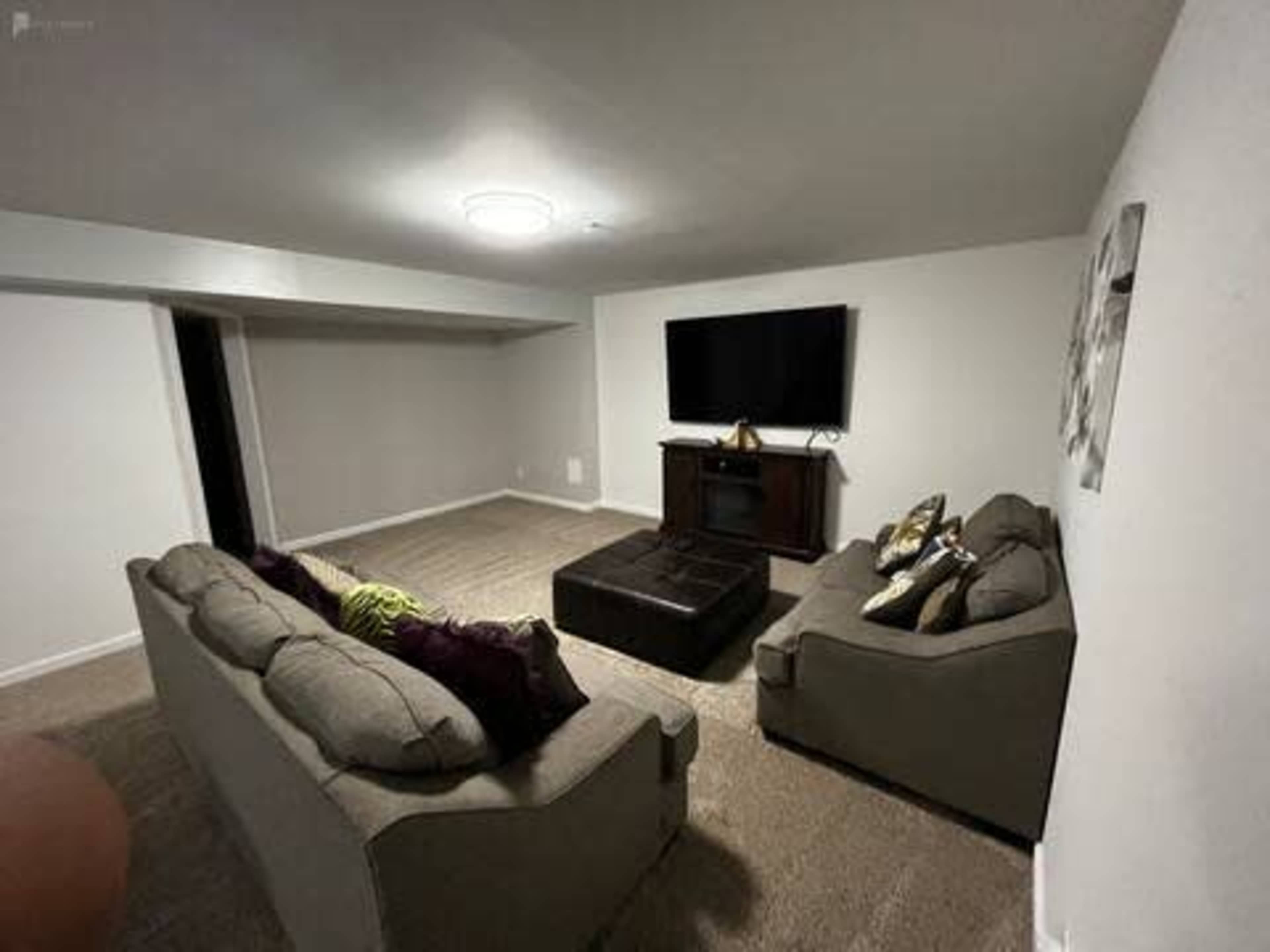 A living room features two sofas, a large ottoman, and a wall-mounted television in a neutral-toned space.