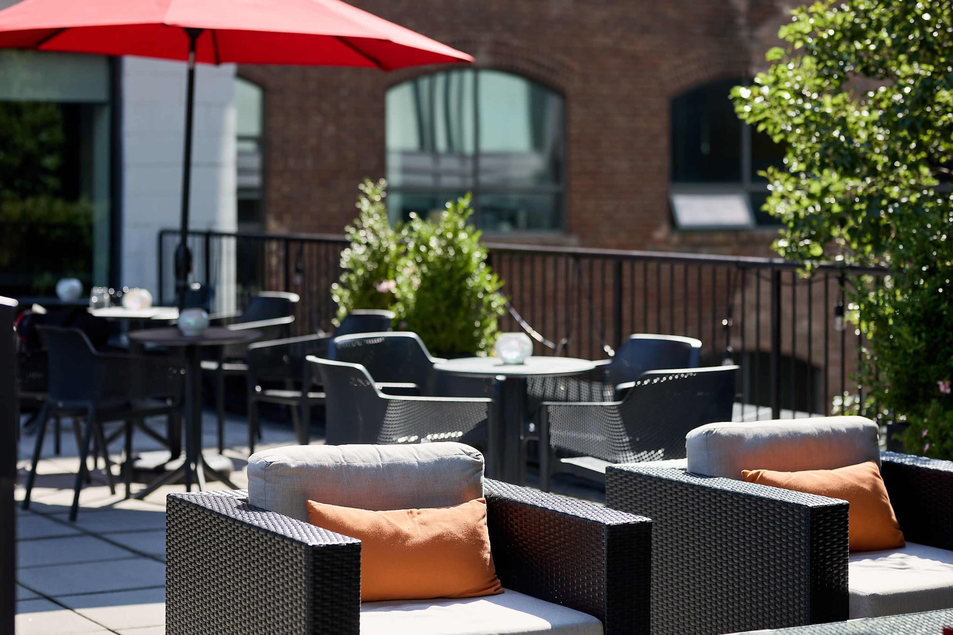 A rooftop patio features modern seating with cushions, a red umbrella, and several tables and chairs against a brick building backdrop.