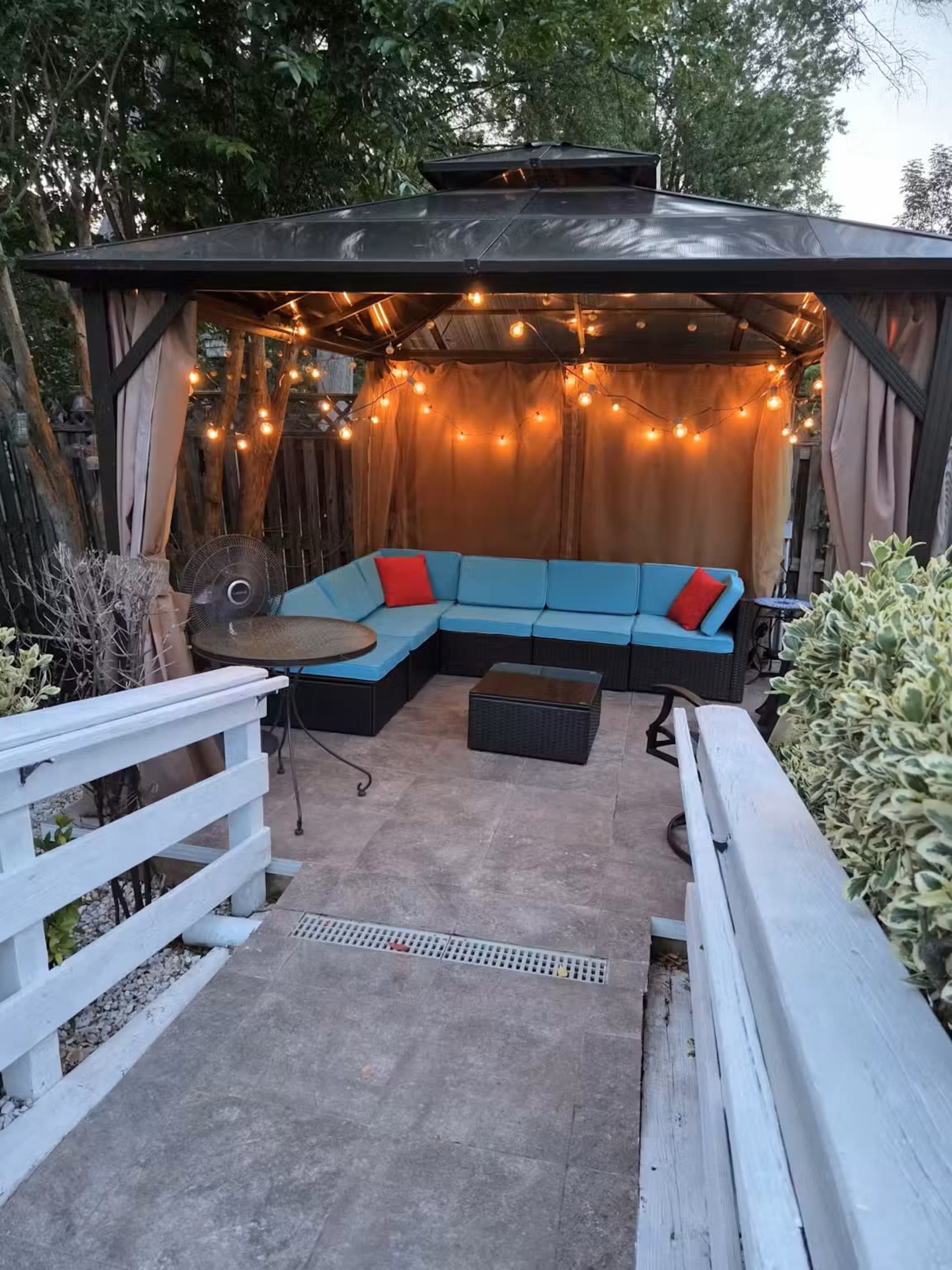 A gazebo with string lights features a corner seating arrangement and a small table, surrounded by greenery.