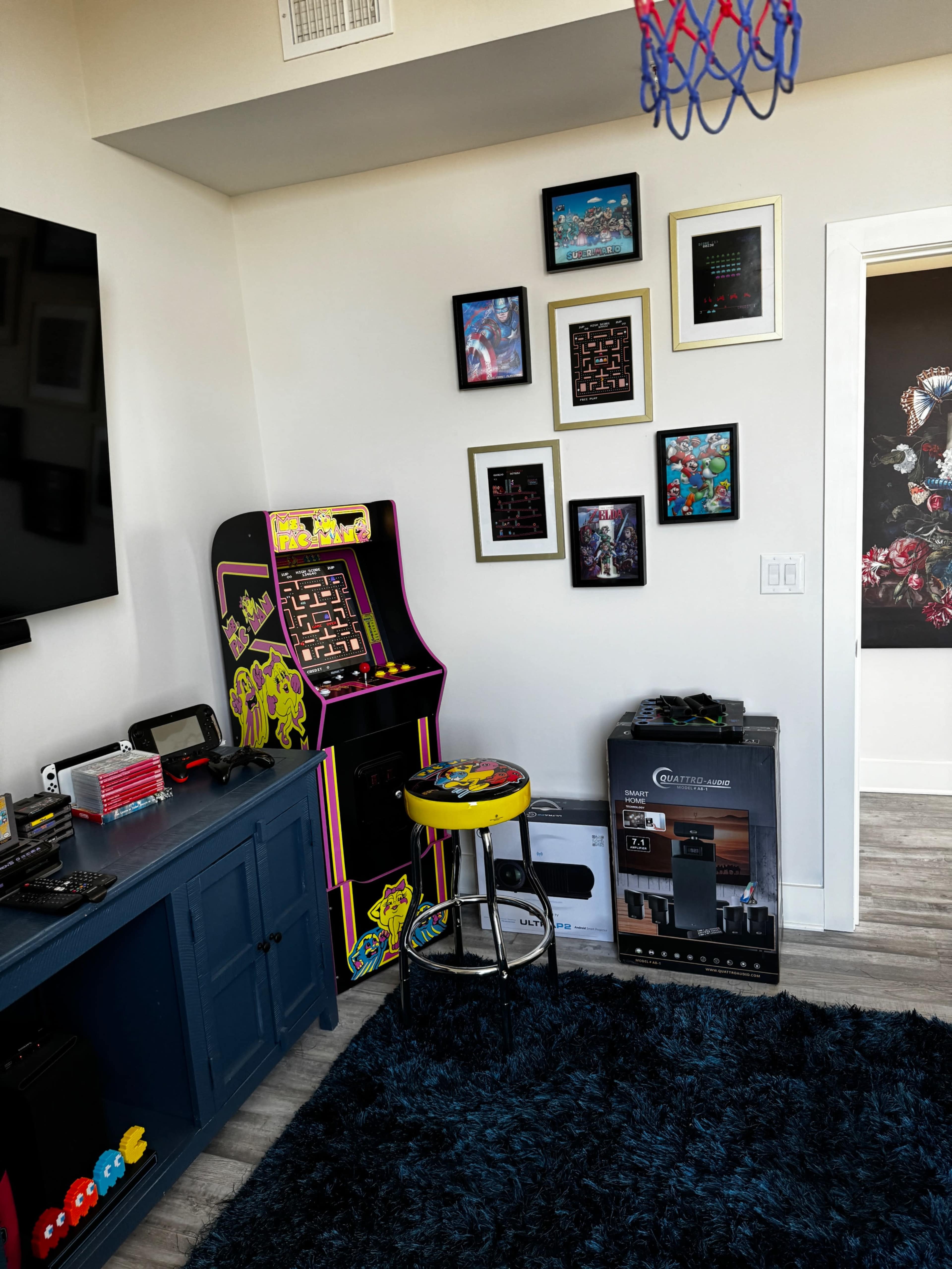 The image shows a room with an arcade game machine, a stool, framed artwork on the walls, and a blue rug.