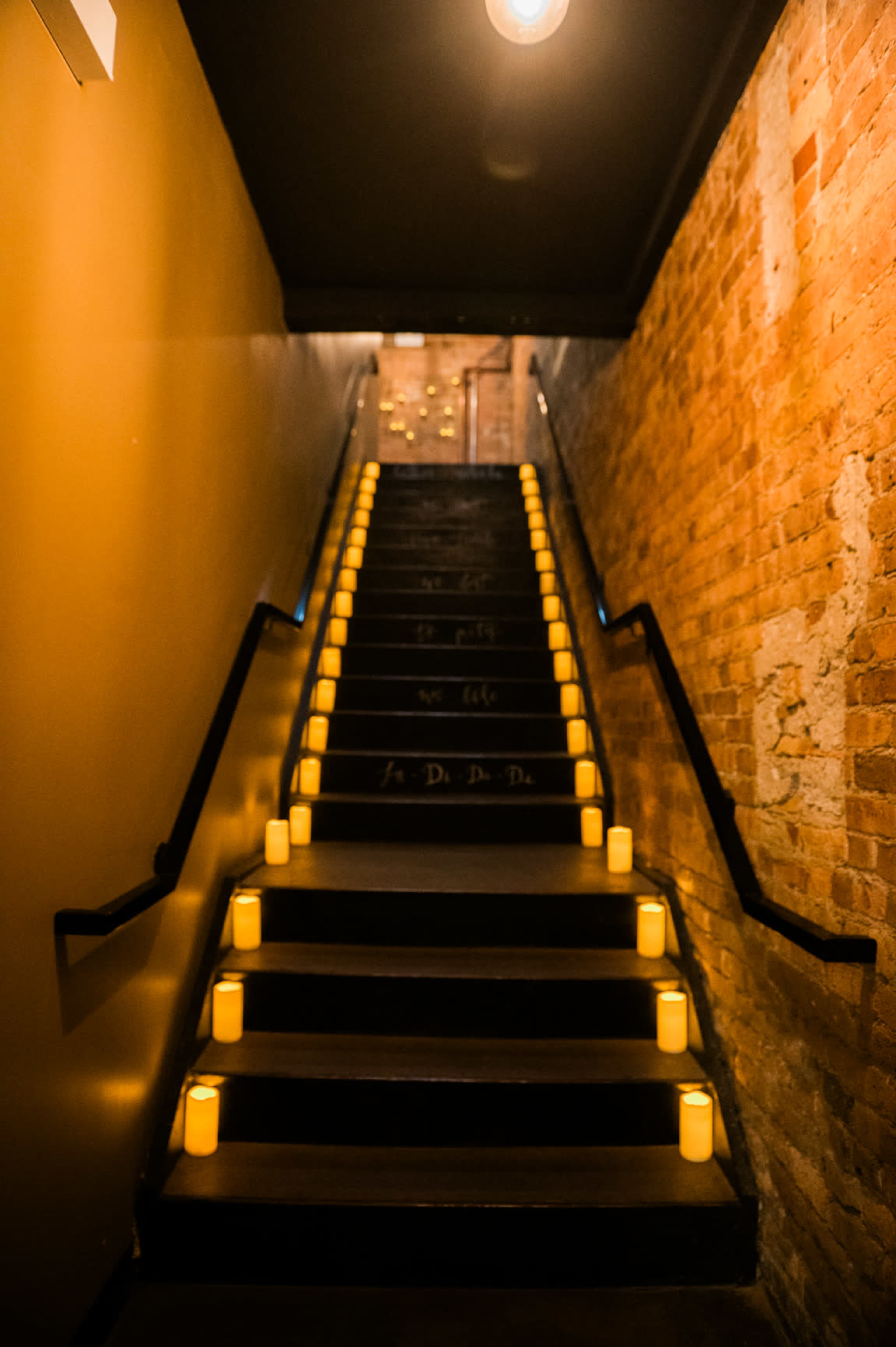 A dimly lit staircase with a brick wall and candles lining each step leads upwards.