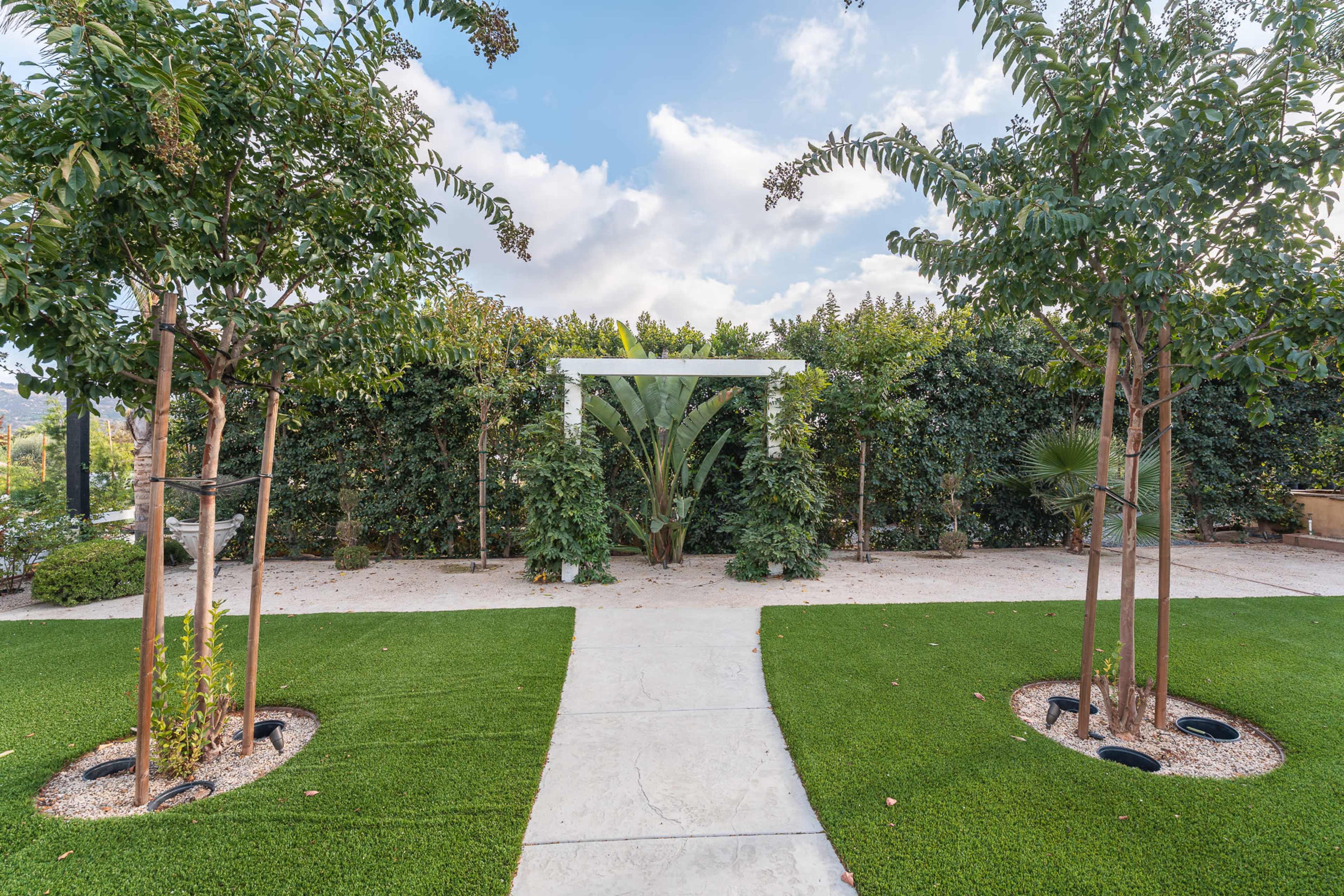 A pathway leads through a landscaped garden featuring neatly trimmed grass, small trees, and a trellis surrounded by lush foliage.