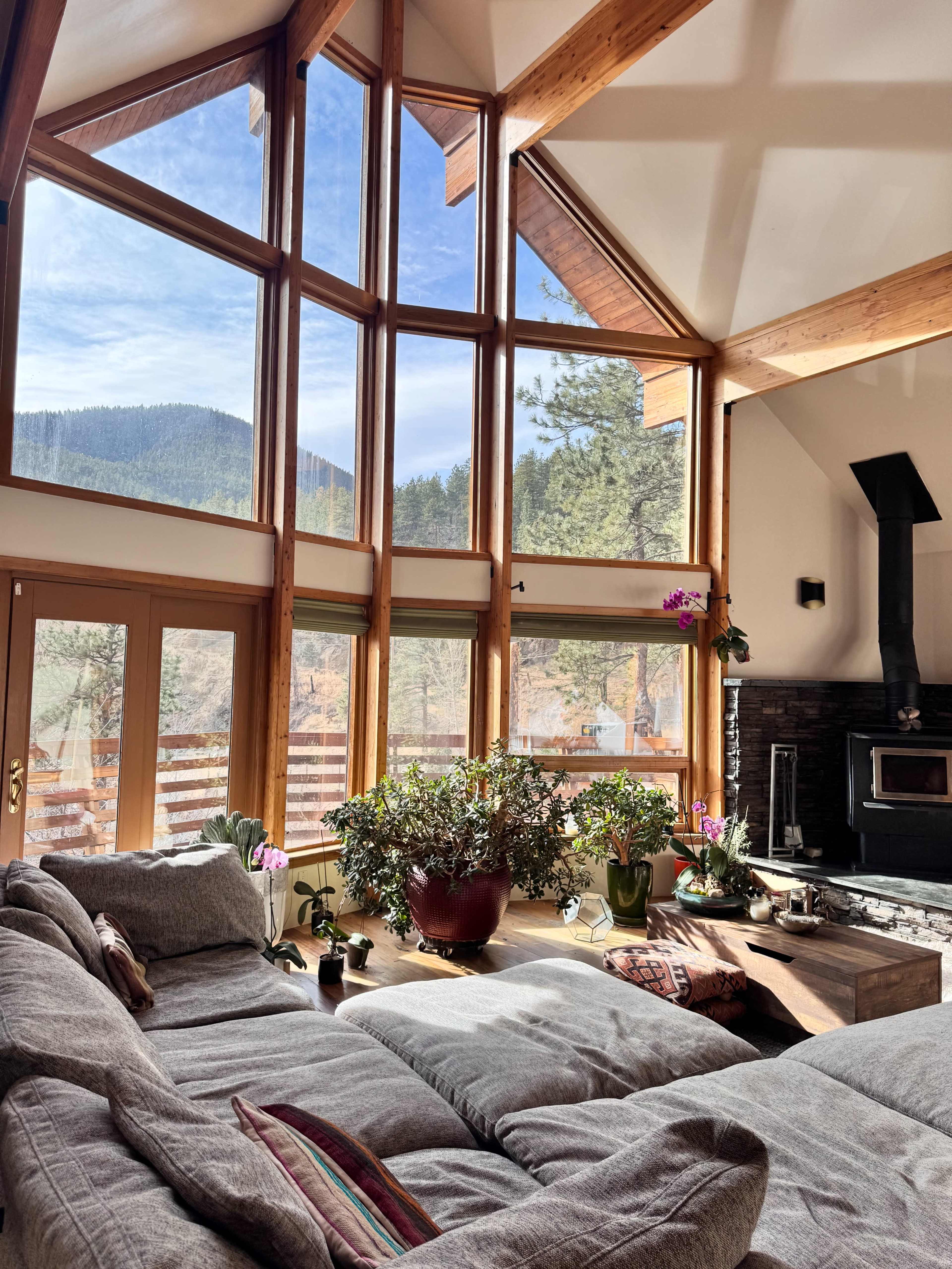 A spacious living room features large windows with mountain views, a cozy seating area, and potted plants.