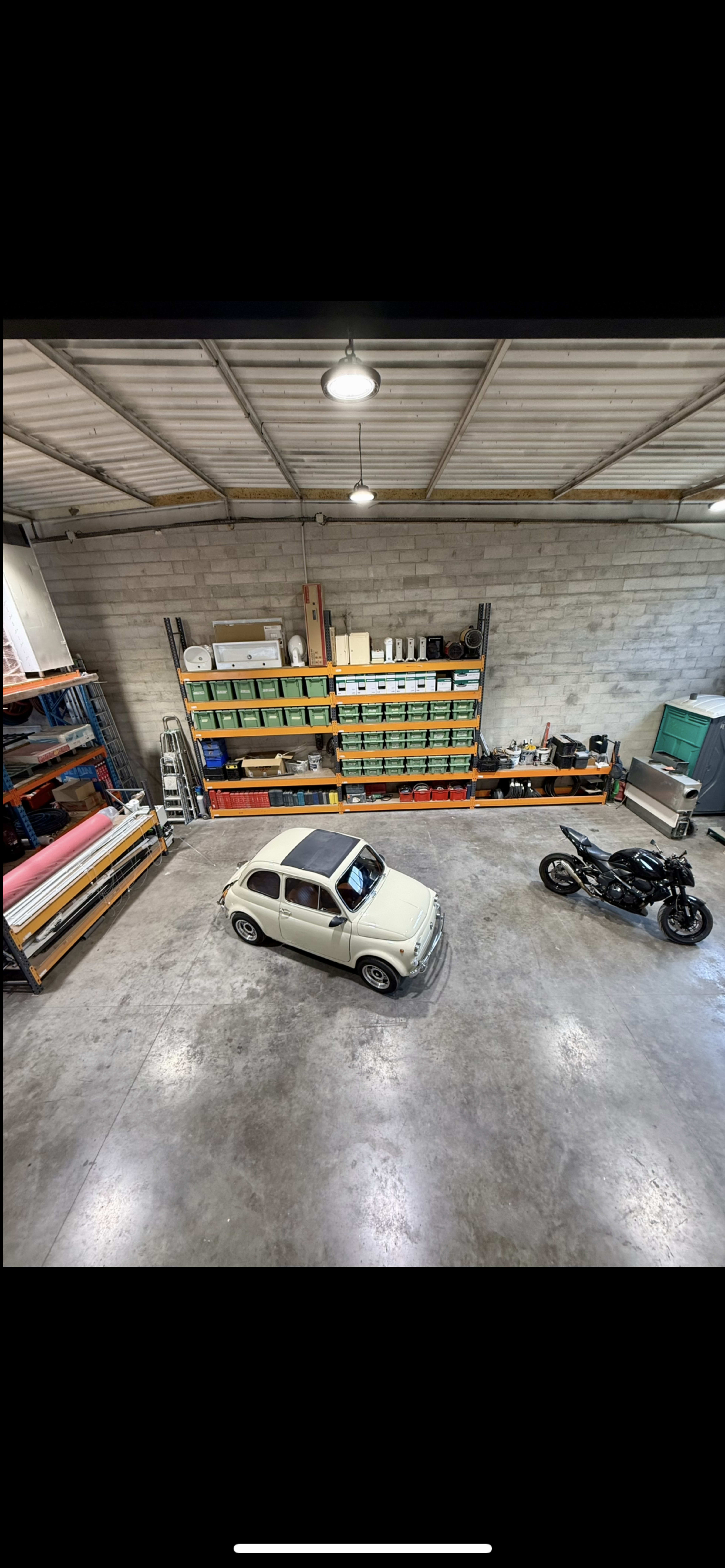 The image shows a spacious garage featuring a small vintage car in the center and a motorcycle on the side, with storage shelves filled with boxes in the background.