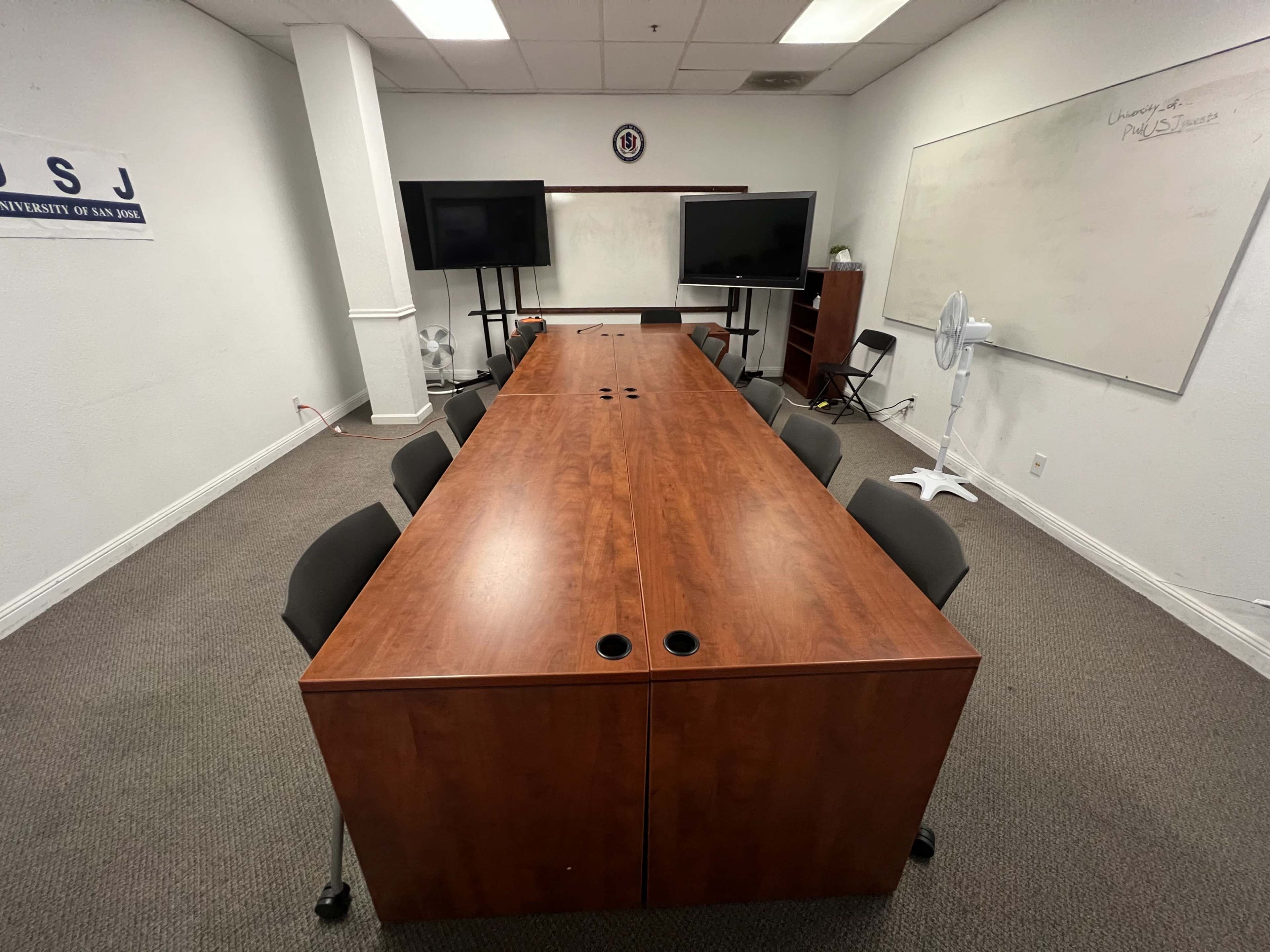 A conference room with a long wooden table surrounded by black chairs, two screens at the front, and a whiteboard on the wall.