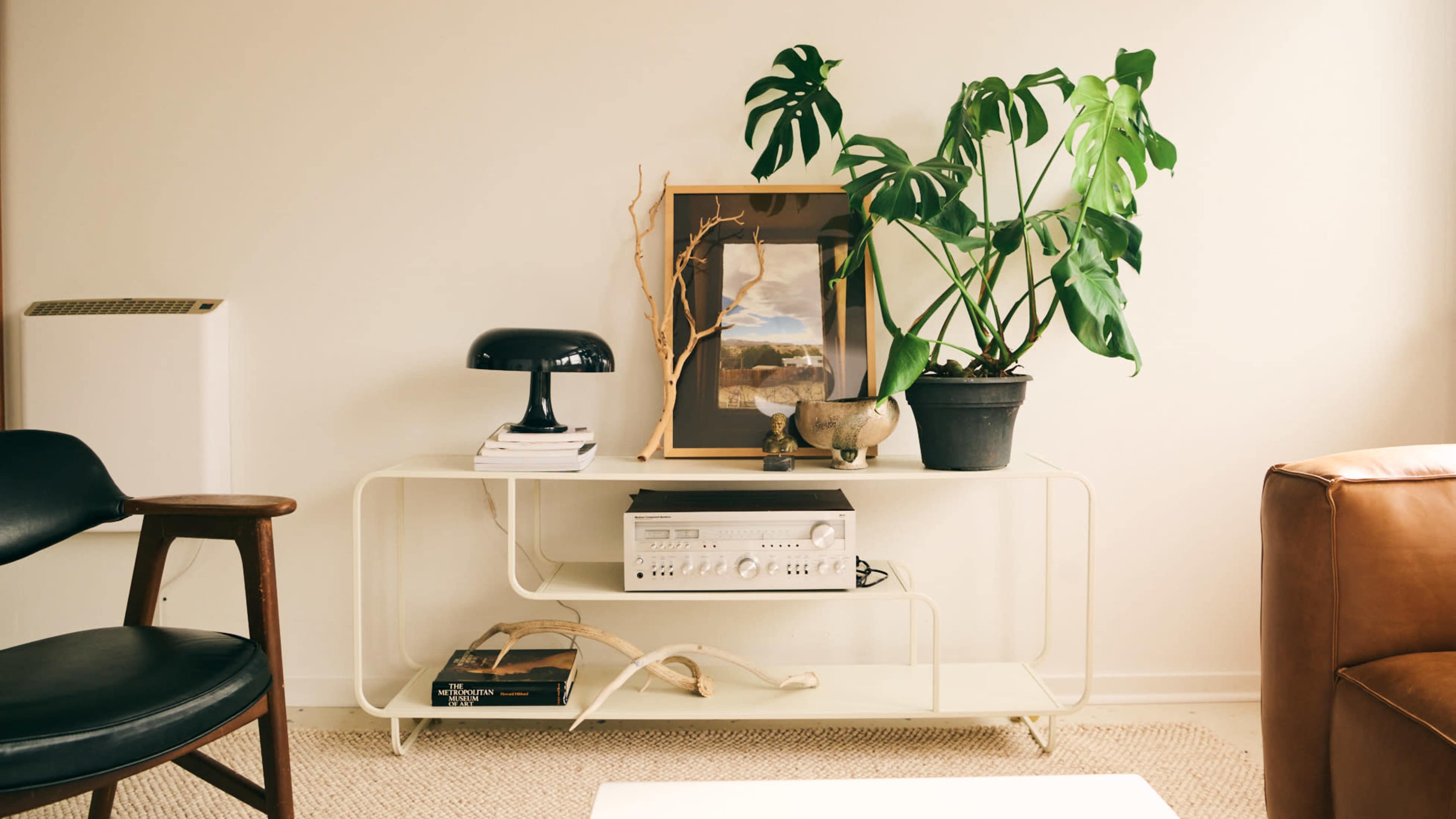 A minimalist living space features a white console table adorned with a planter, framed artwork, a lamp, and a stack of books, next to a black chair and a brown leather sofa.