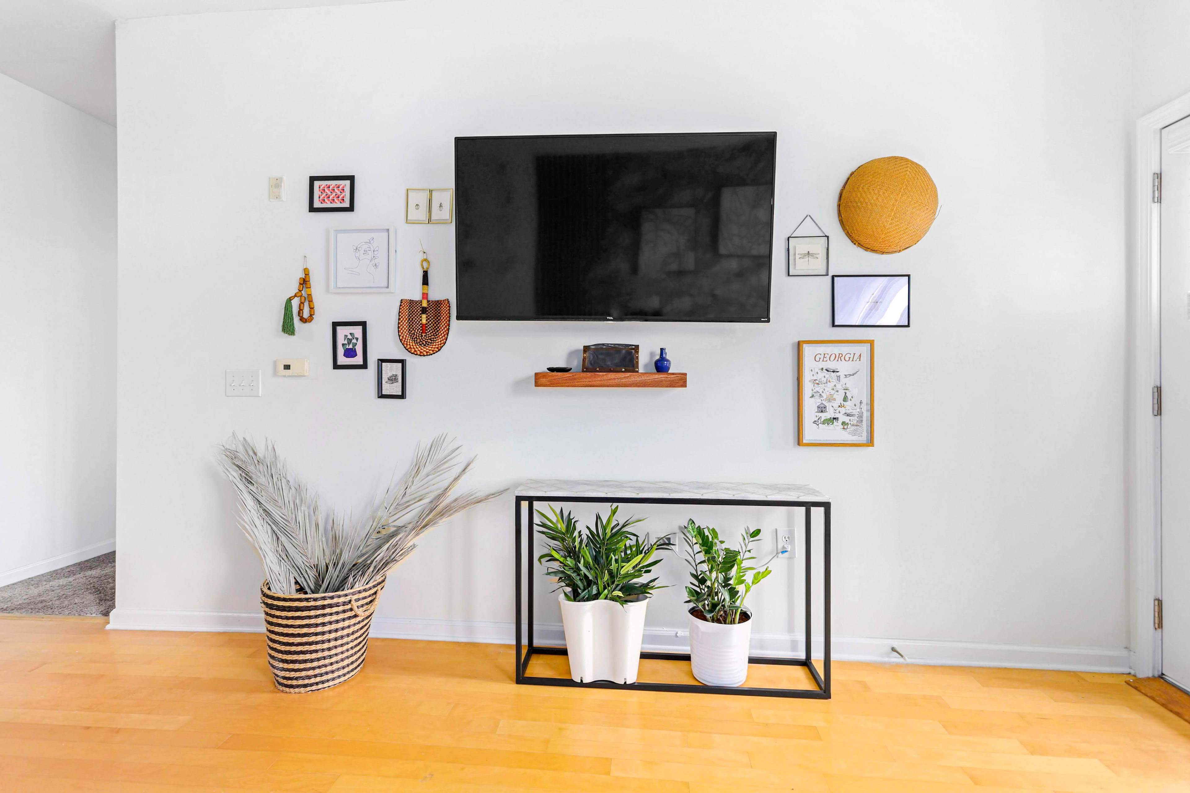A modern living room wall features a mounted flat-screen TV above a console table, adorned with plants and framed artwork.