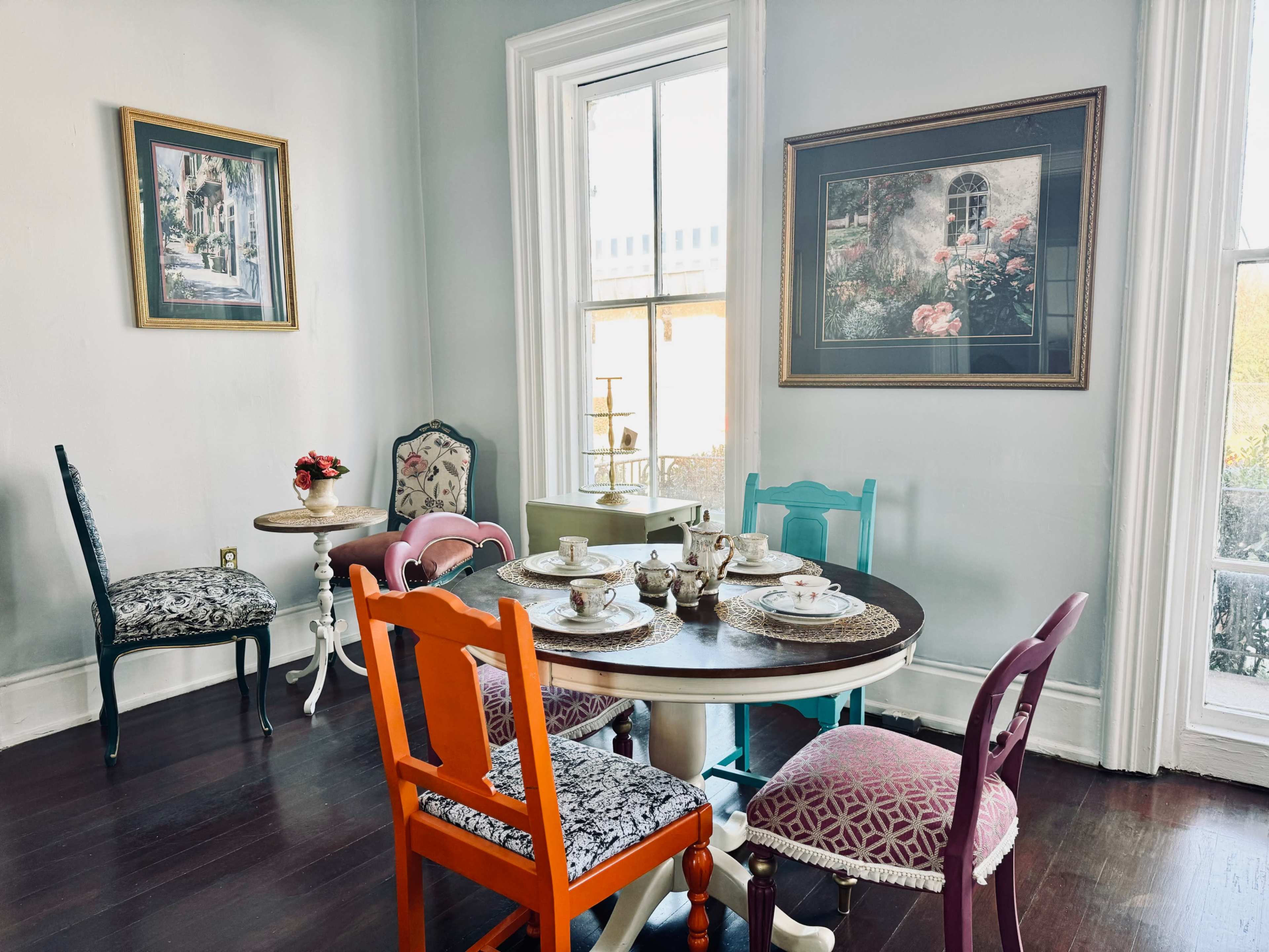 A dining area features a round table set with tea cups and a variety of colorful chairs, along with framed artwork on the walls and a window allowing natural light.
